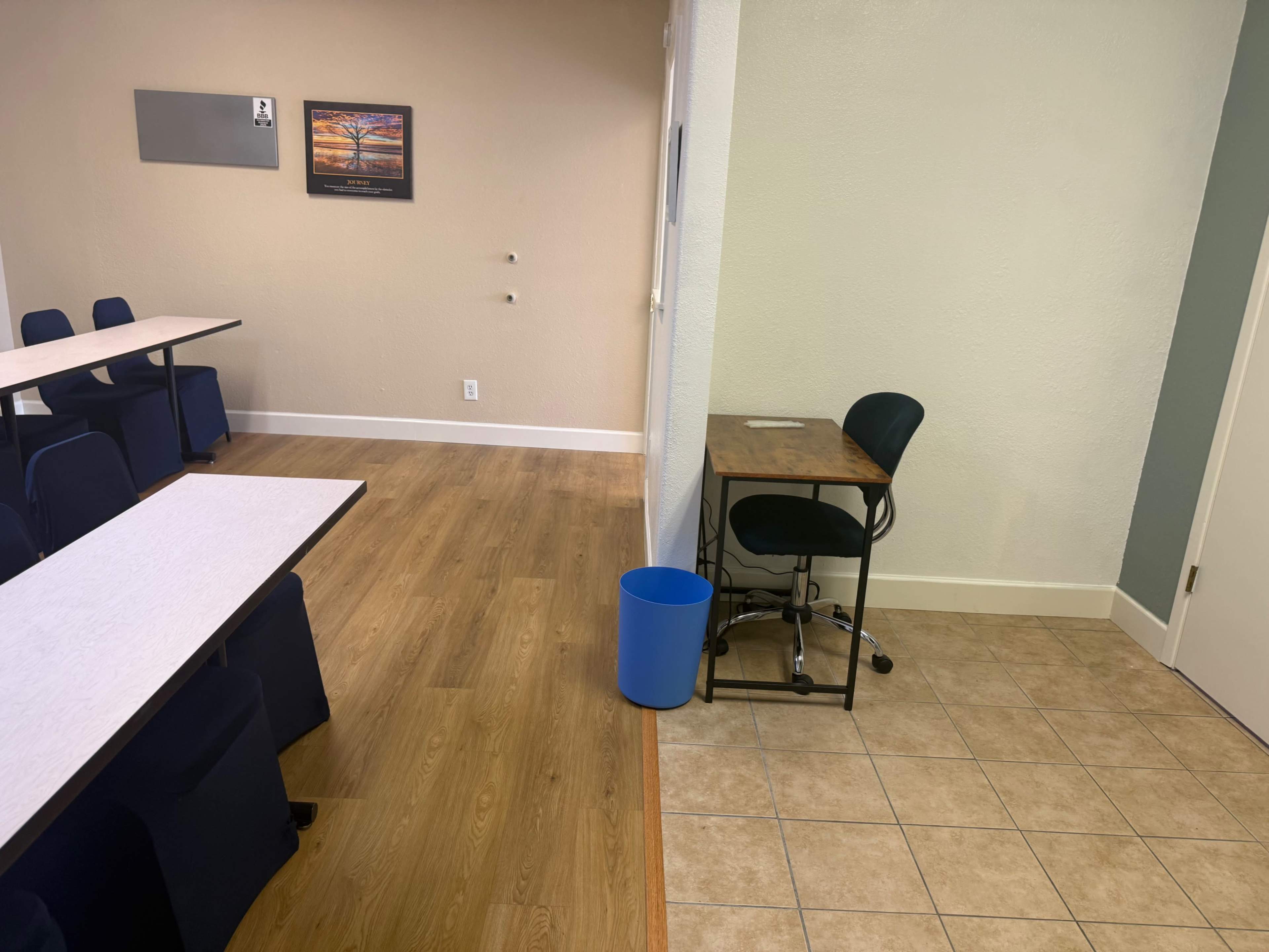 The image shows a split room with tables and chairs on the left side and a desk with a chair next to a blue trash can on the right side.