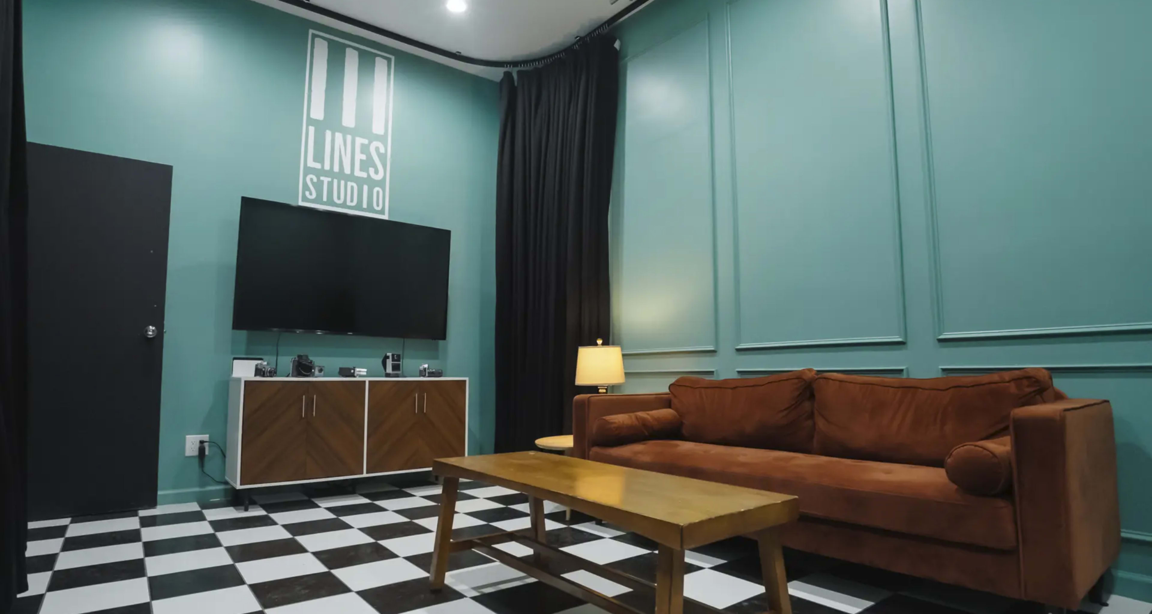 The image shows a modern studio room with teal walls, a brown couch, a wooden coffee table, and a television mounted on the wall.