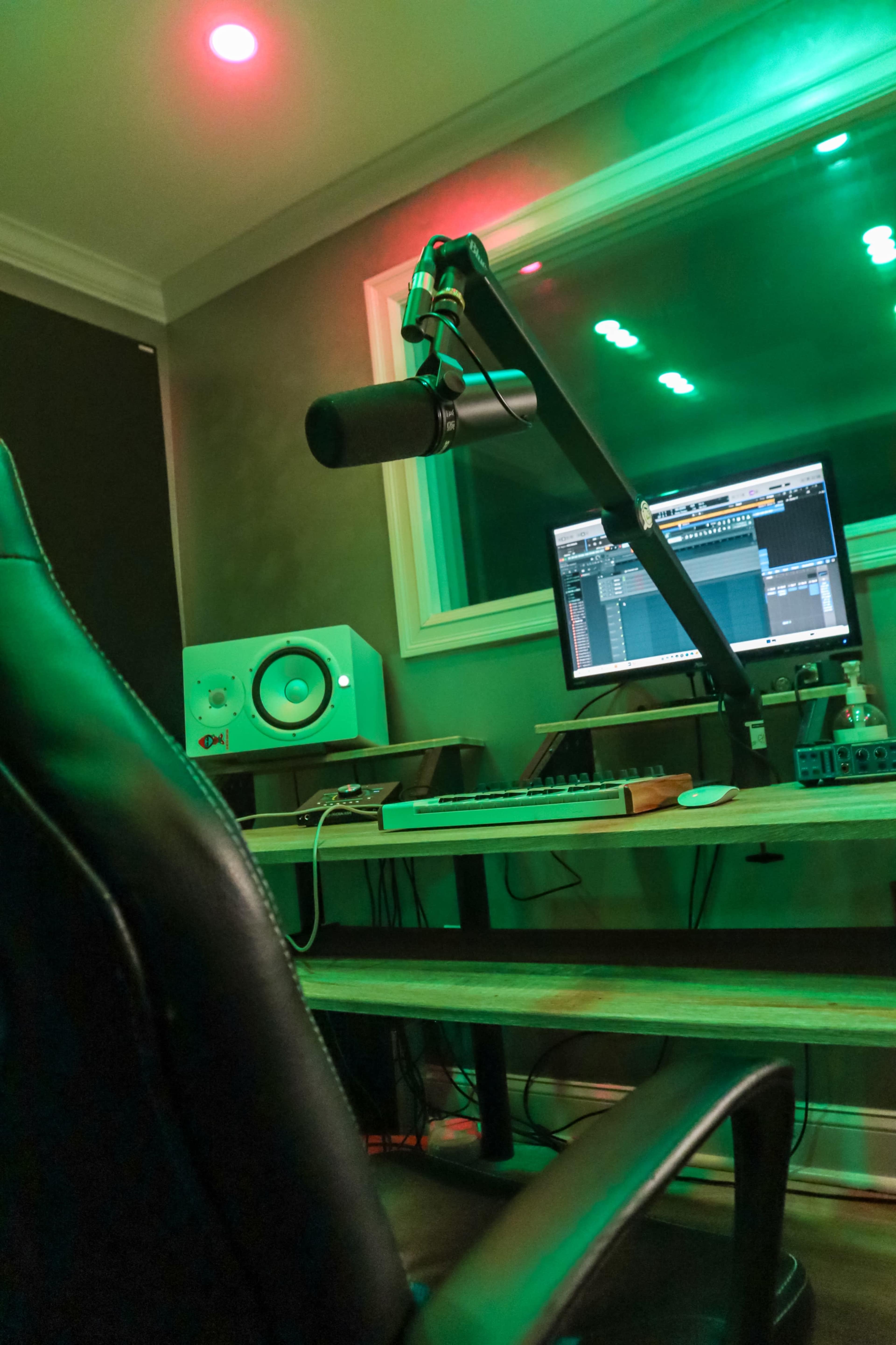 A microphone is mounted beside a computer with music production software in a recording studio setting with green and red lighting.