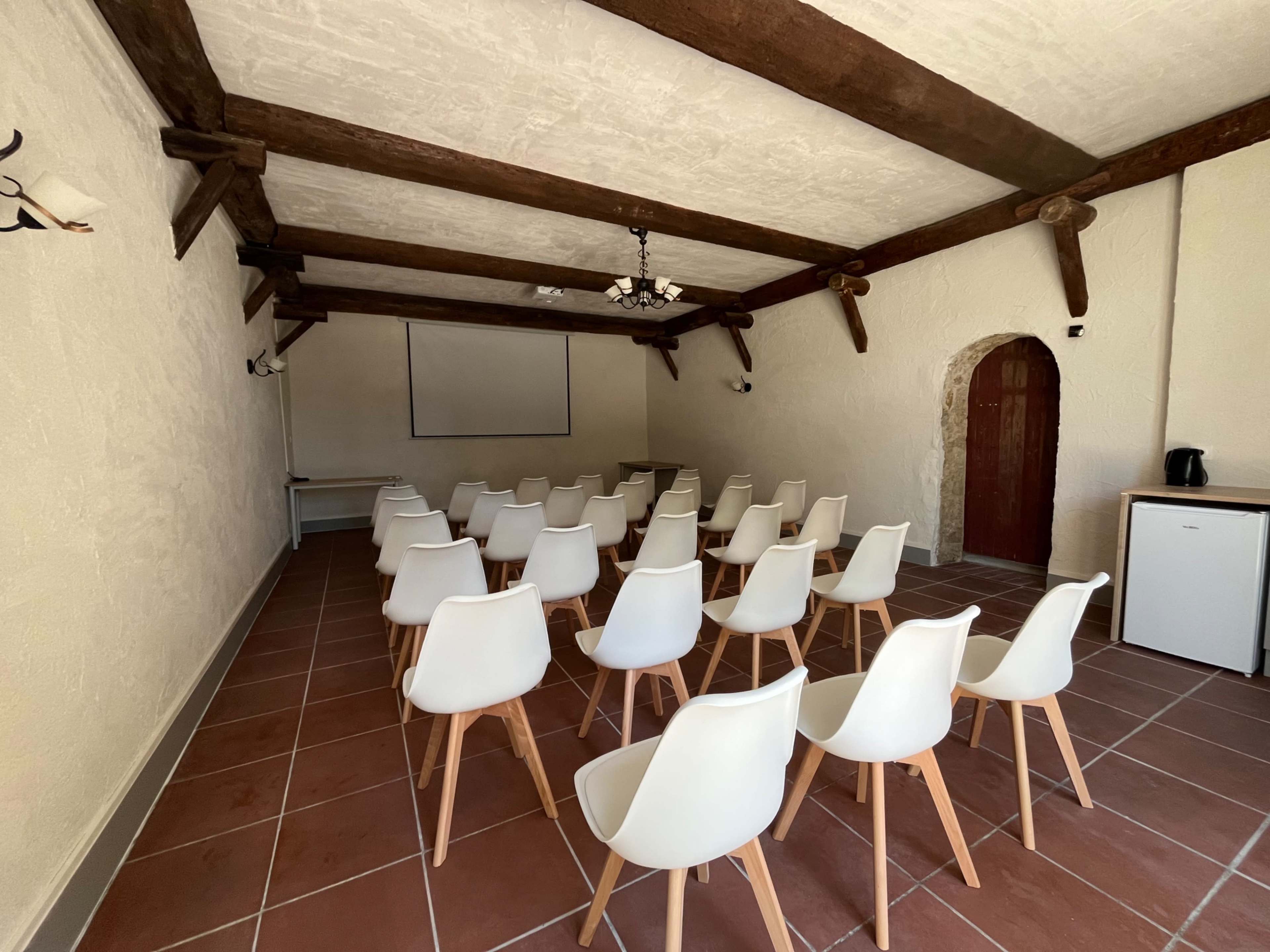 The image shows a meeting room with rows of white chairs arranged facing a blank projection screen against a light-colored wall.