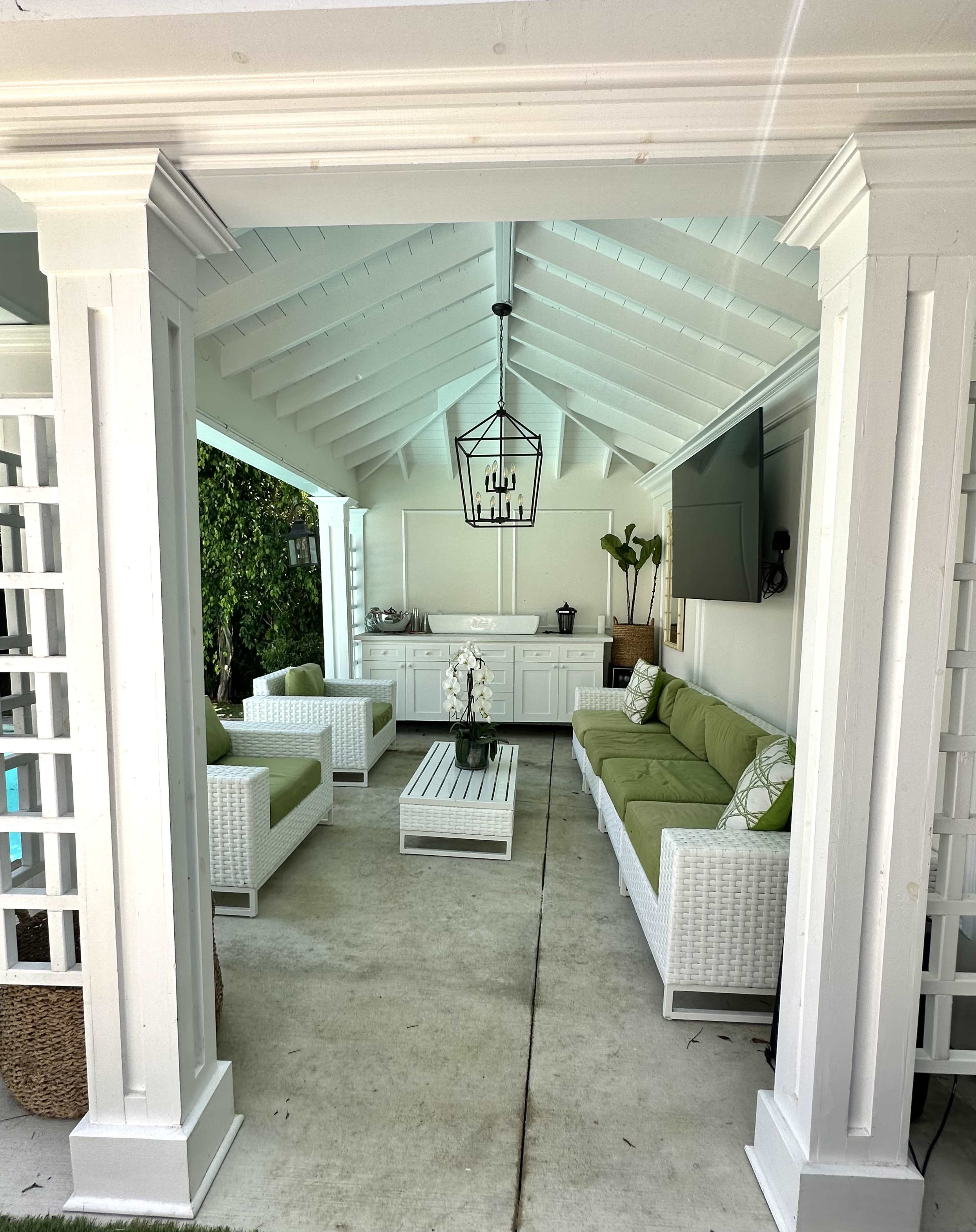 The image shows a covered outdoor lounge area with white wicker furniture and green cushions, featuring a central coffee table and a flat-screen TV mounted on the wall.