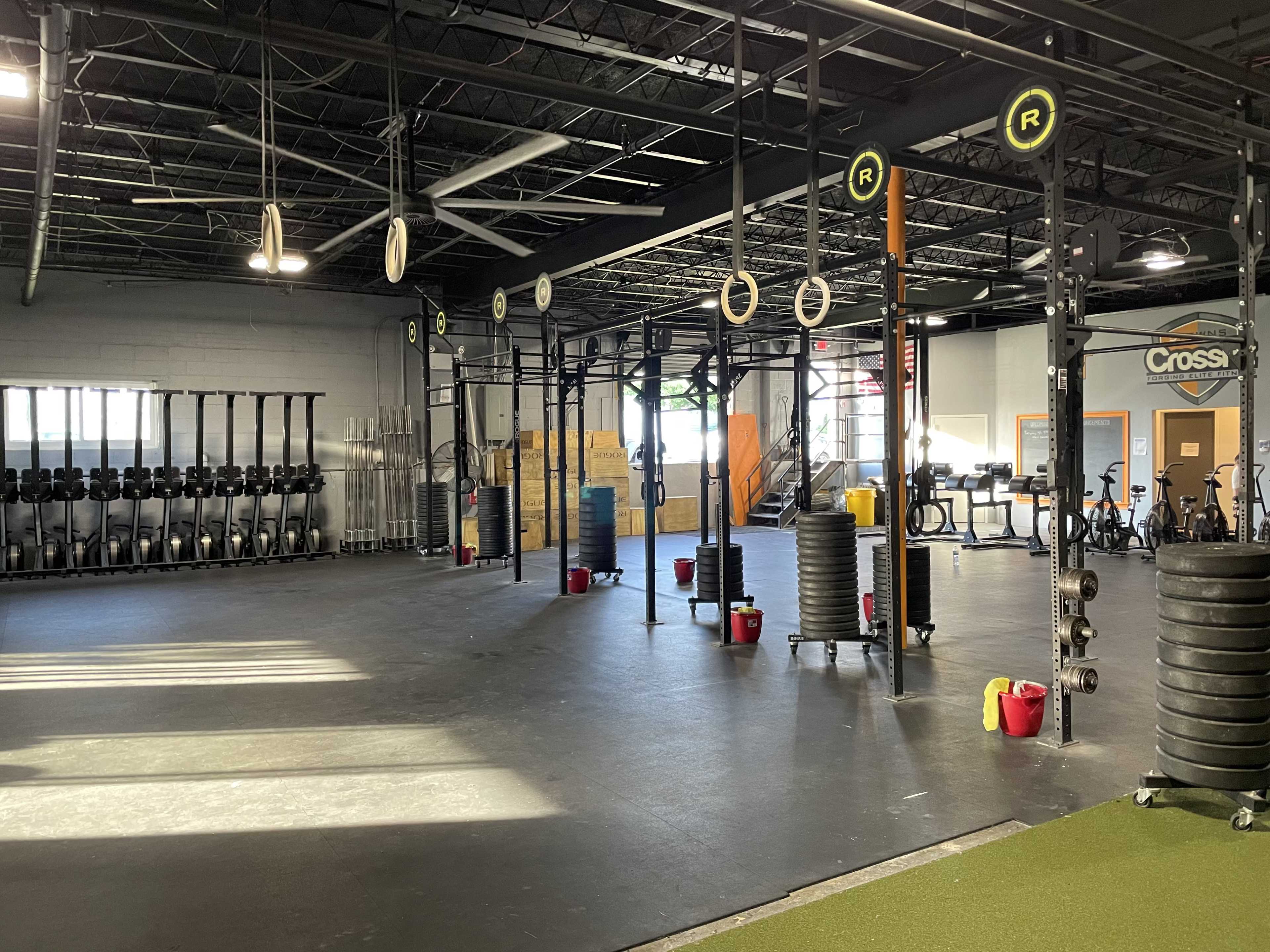 The image shows a spacious gym area with weightlifting equipment, exercise mats, and a row of rowing machines along the wall.