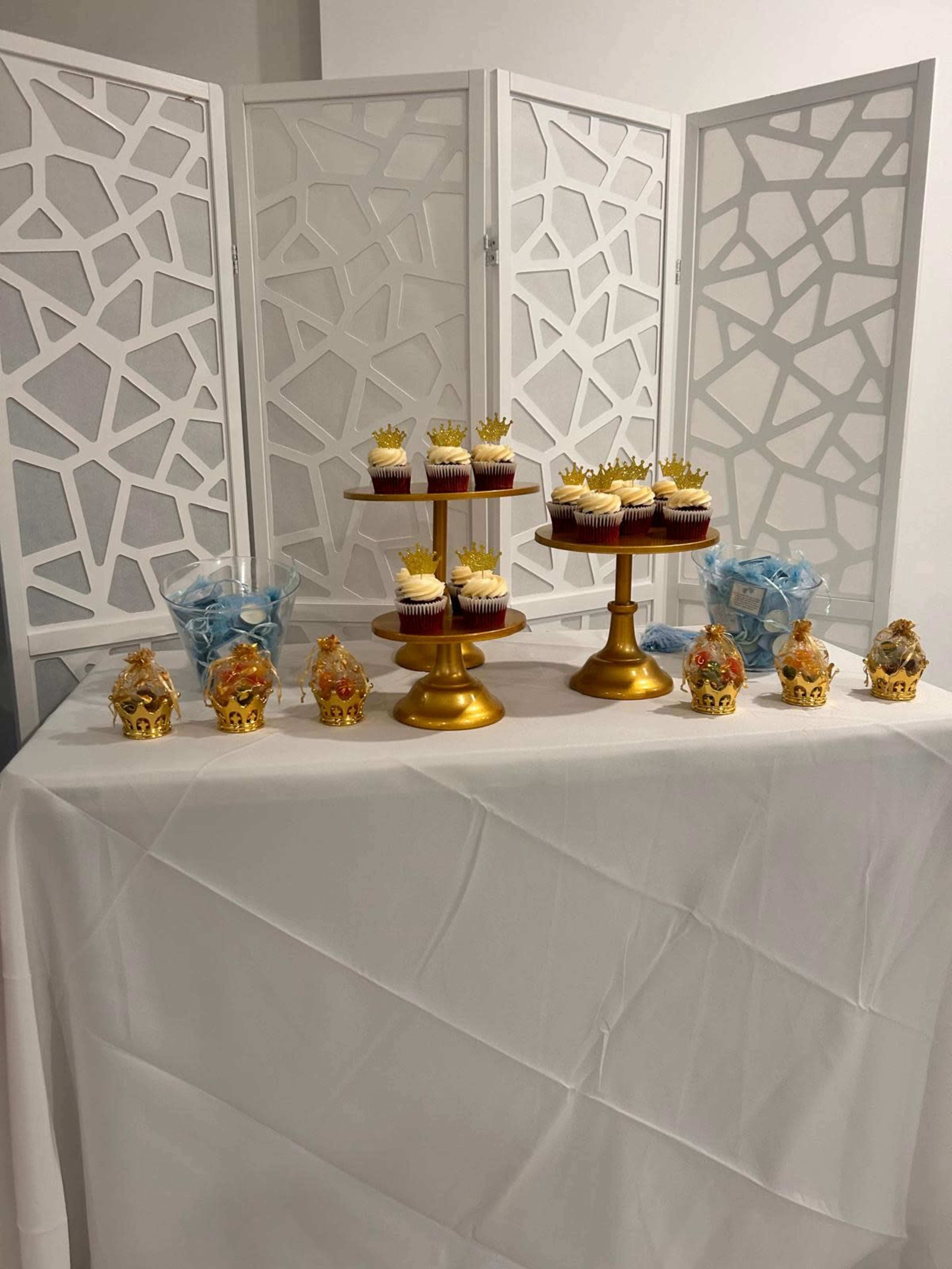 The image shows a dessert table featuring multiple tiers of cupcakes on gold stands, with decorative blue accents and ornate figurines arranged on a white tablecloth.