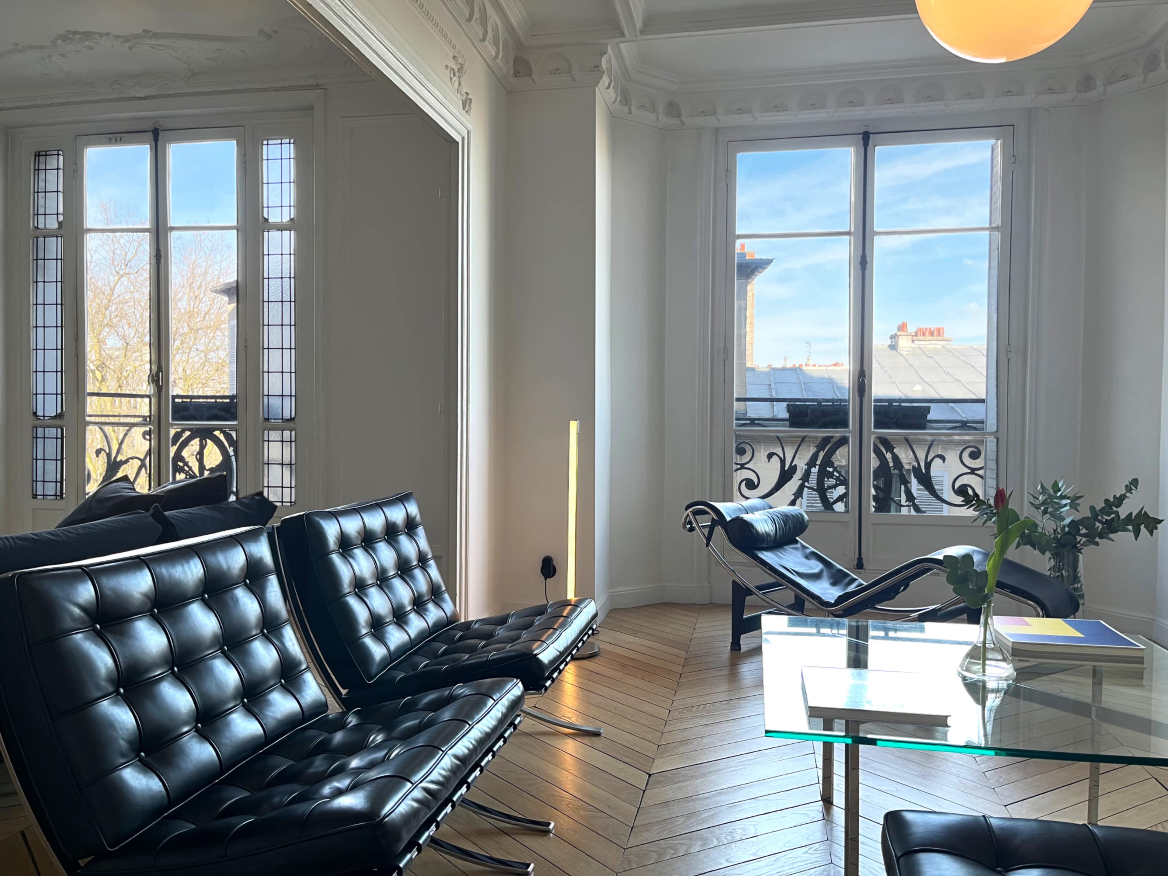 The image shows a stylish living room with black leather furniture, a glass table, and large windows that provide a view of a blue sky outside.