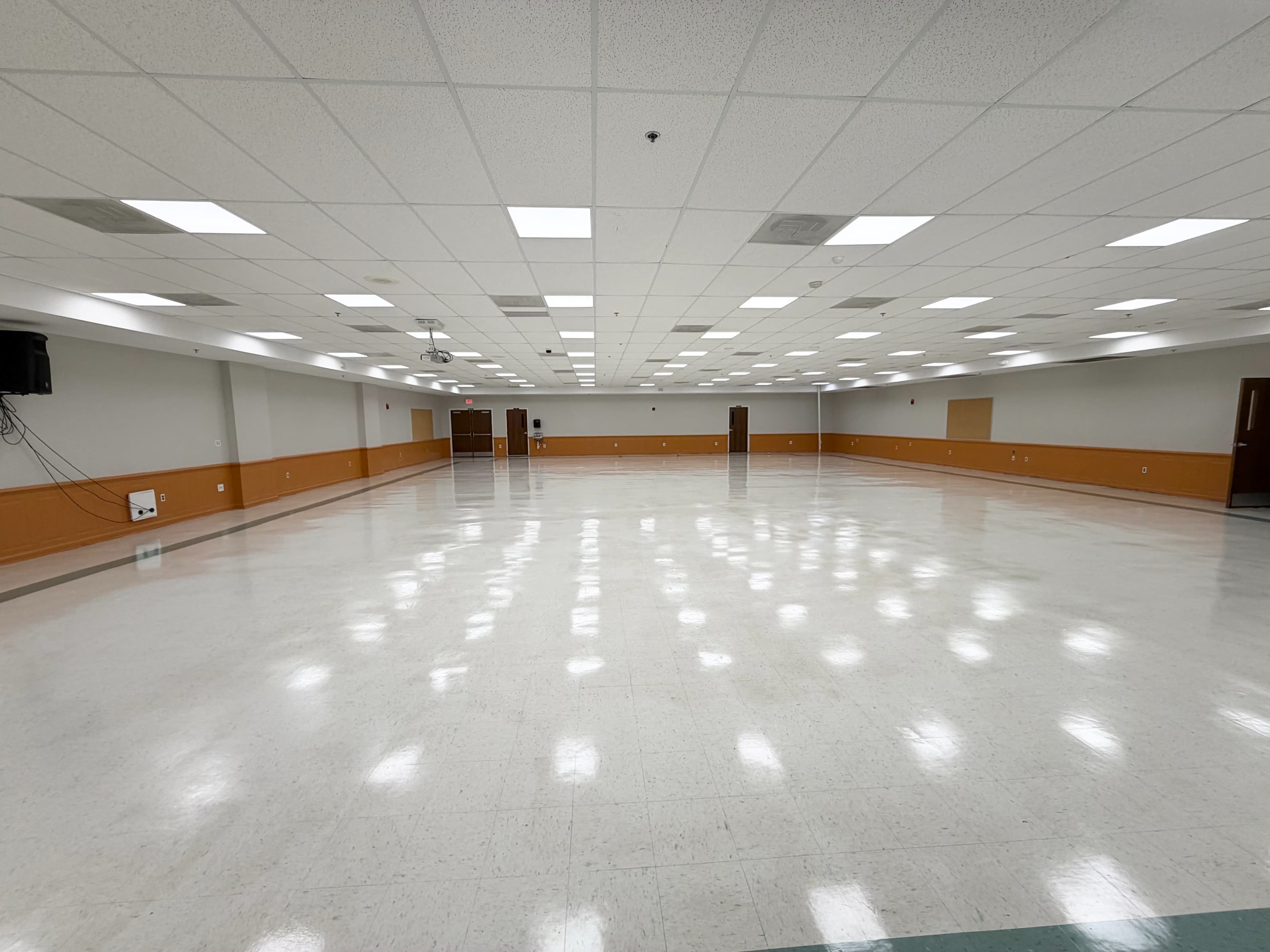 The image shows a large, empty room with white tiled floors and orange-painted walls, featuring fluorescent lighting and multiple doors along the perimeter.