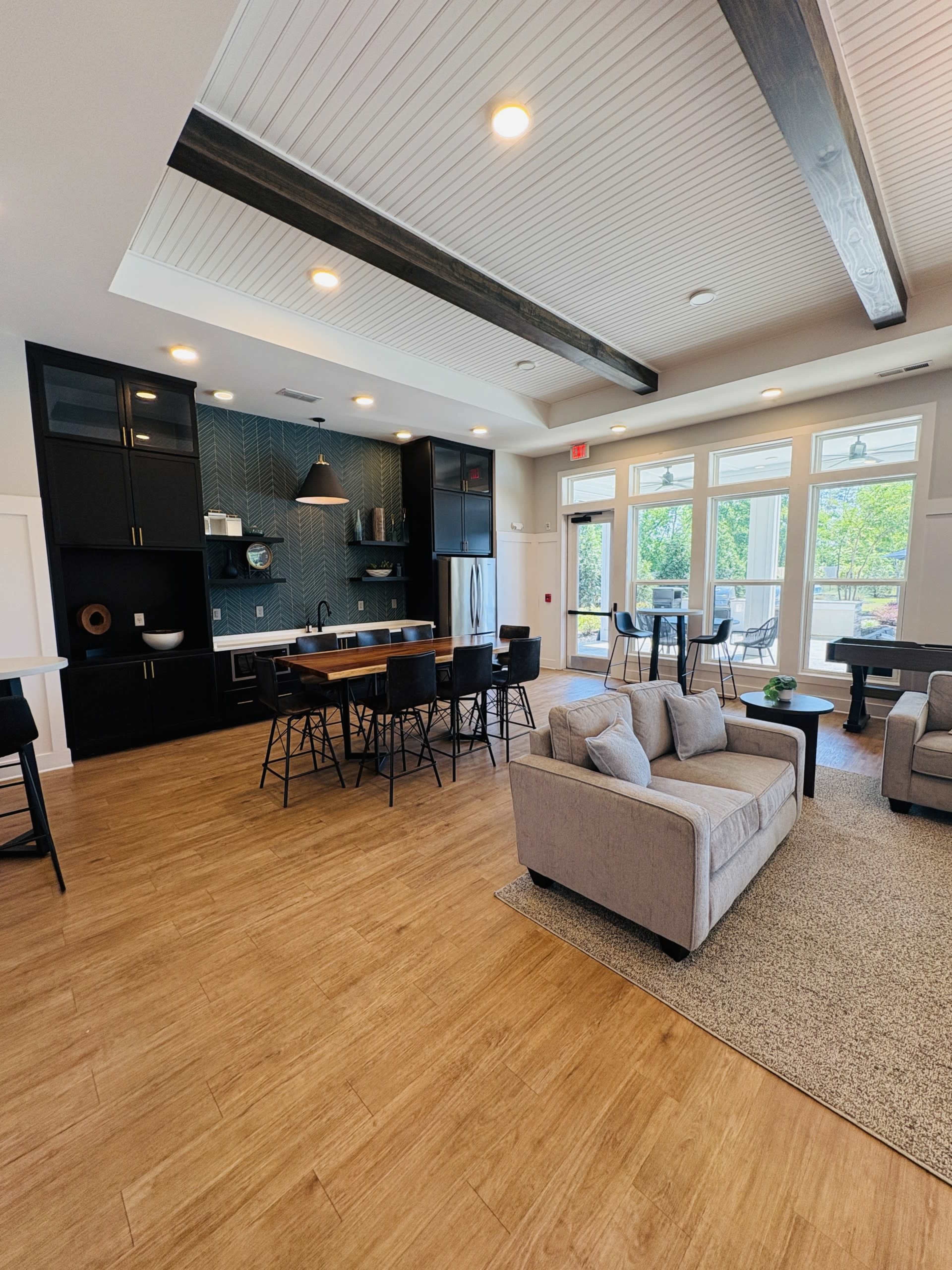 The image shows a modern communal room featuring a kitchen area, dining table, and lounge seating with large windows allowing natural light.