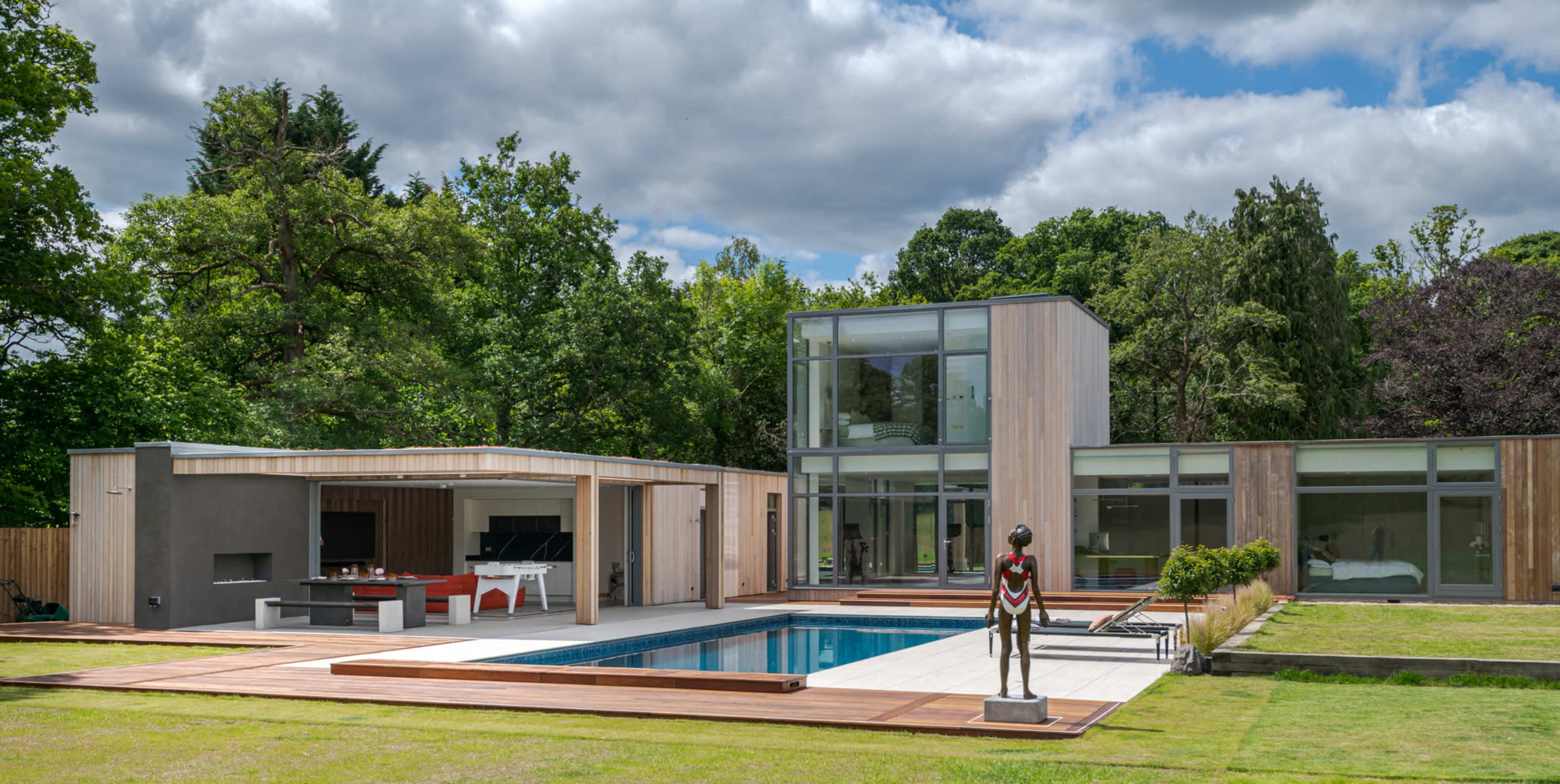 The image shows a modern house with large glass windows, a swimming pool, and a sculpture in the foreground, all set in a landscaped area with trees.