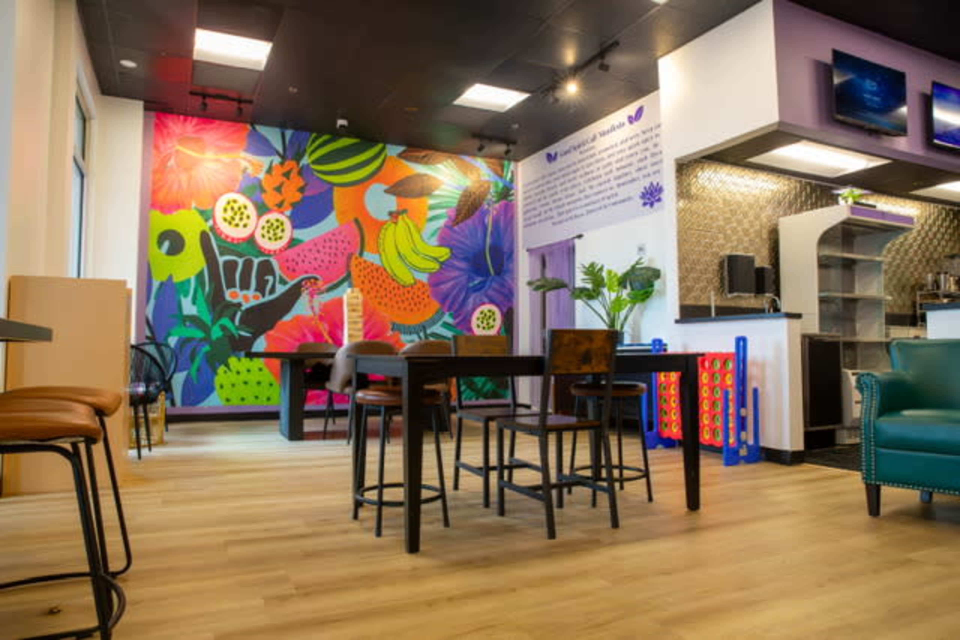 The image shows a brightly colored cafe interior with a large mural featuring fruits and plants, surrounded by various seating arrangements and a service area.