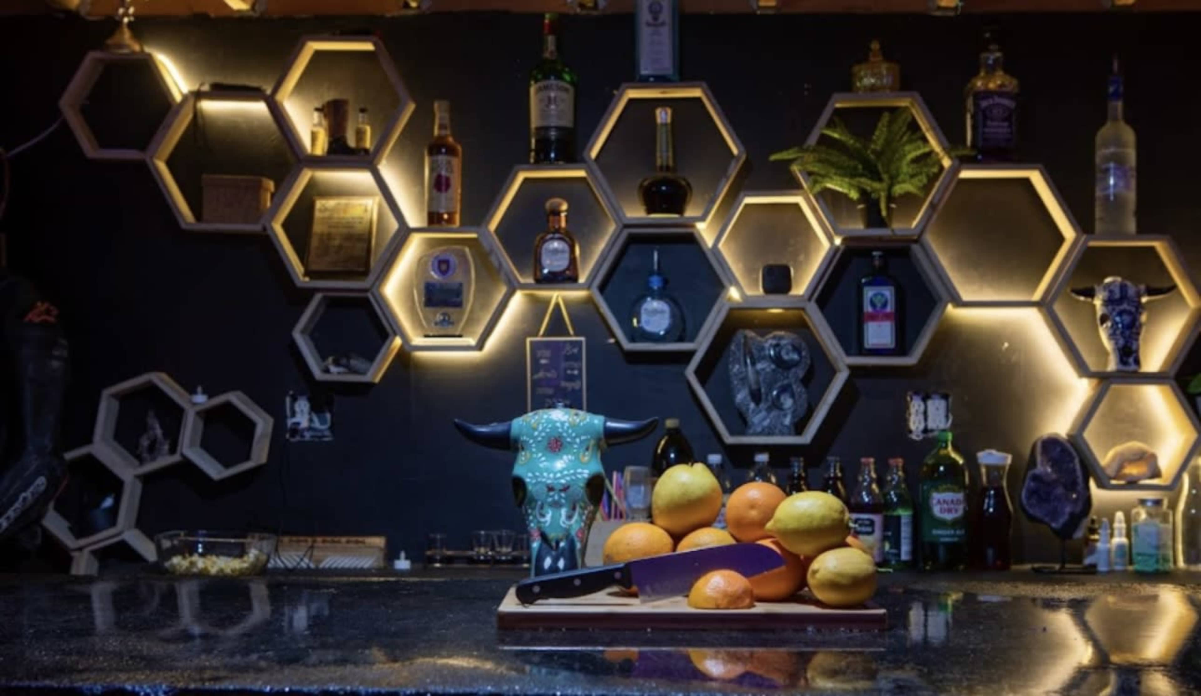 A bar counter with a decorative arrangement of hexagonal shelves displaying various bottles and a cutting board with several fruits in front.