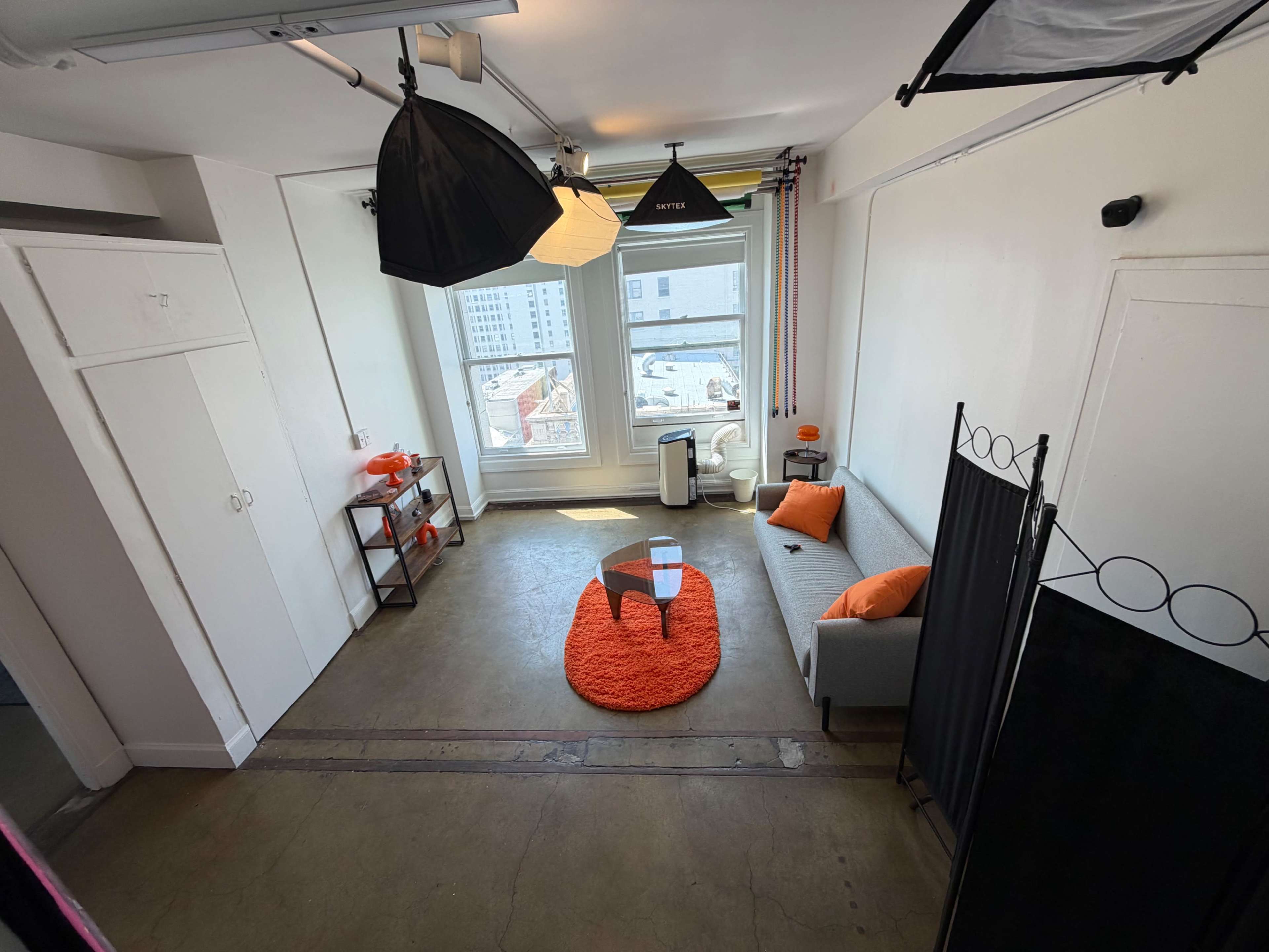 The image shows a modern studio space featuring a gray sofa, an orange rug, a glass coffee table, and two large windows with city views.