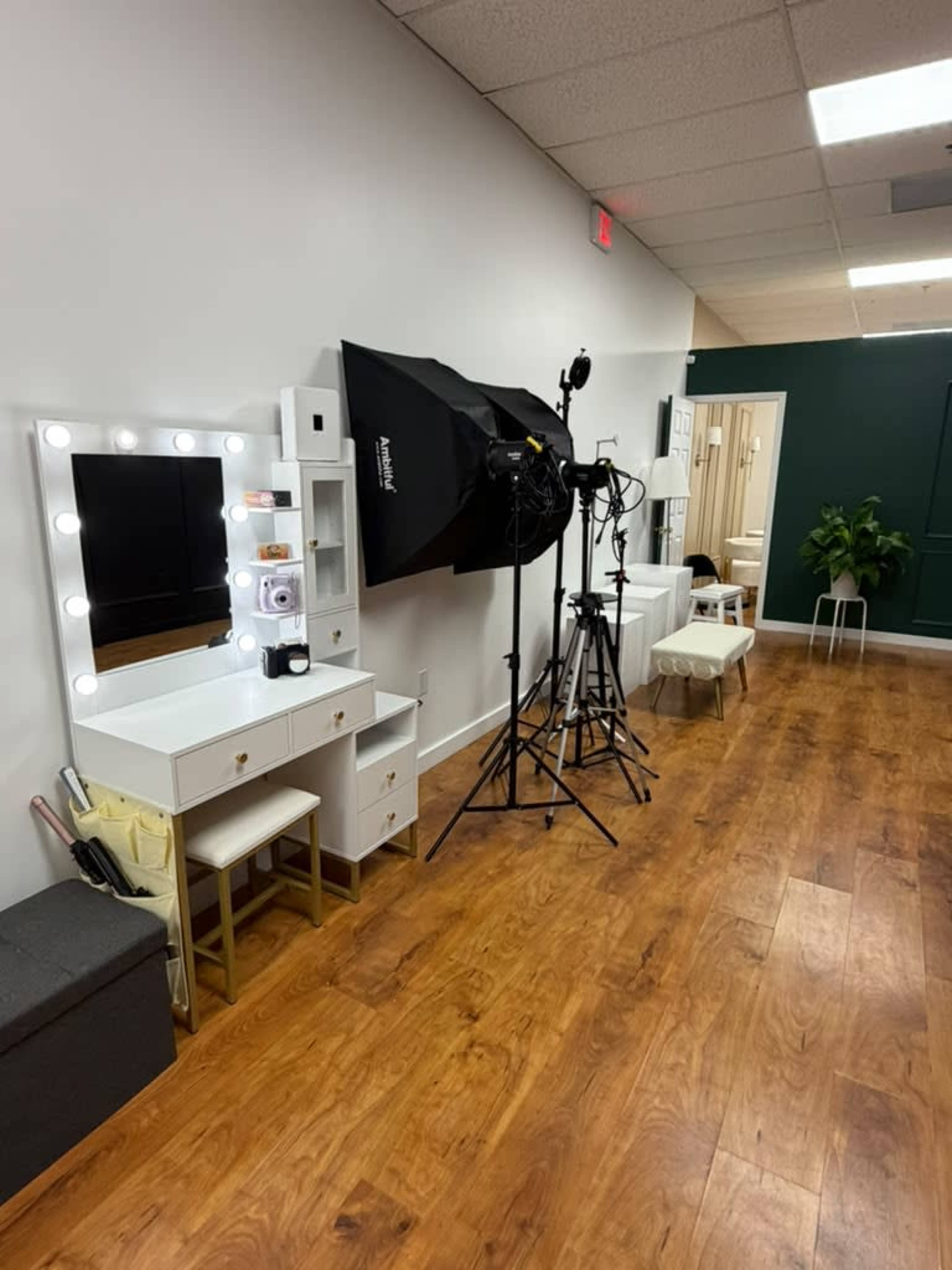 The image shows a well-lit room featuring a vanity mirror with lights, a black photography umbrella softbox, and several tripods set up against a wall with wooden flooring.
