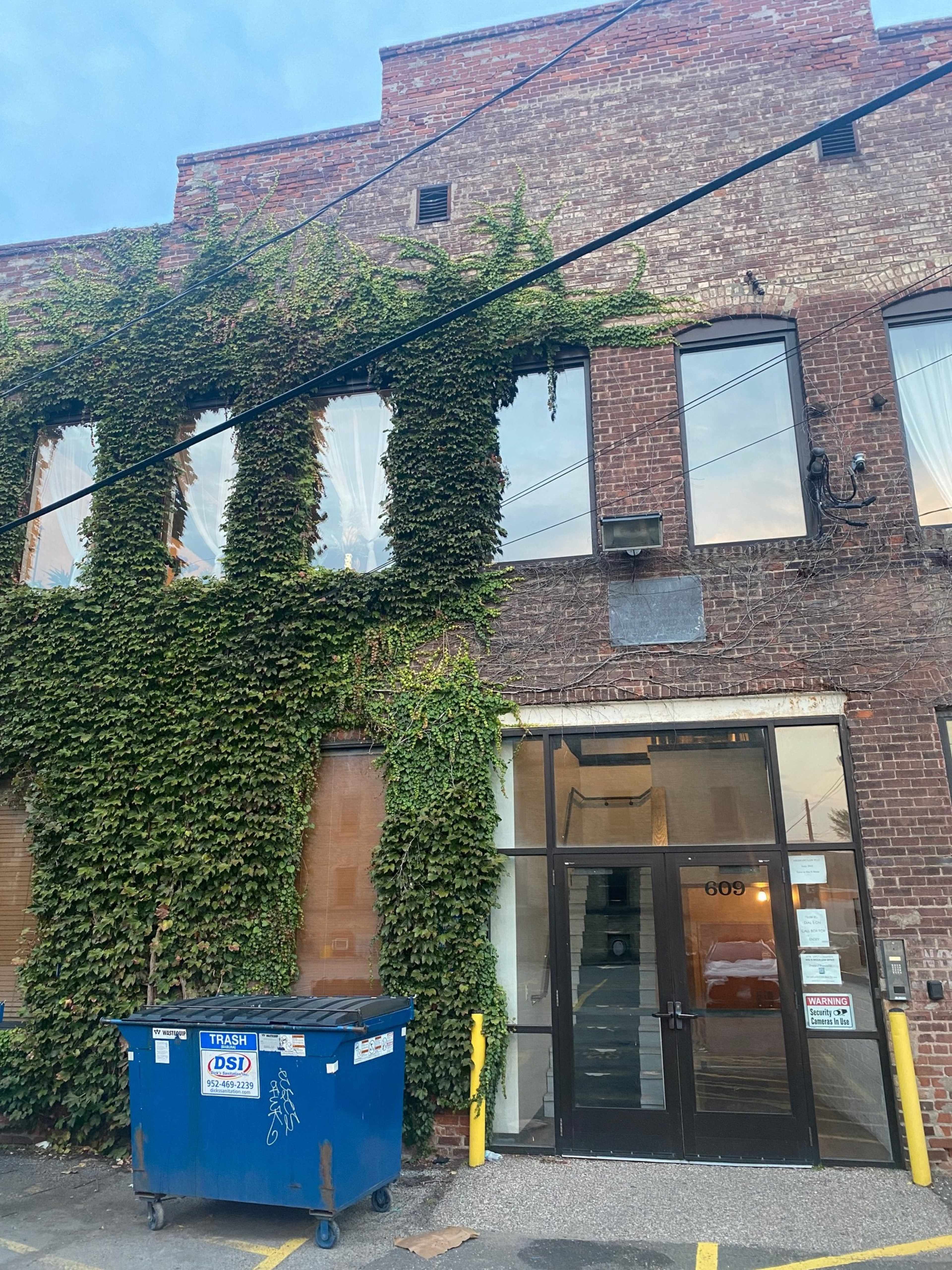 A brick building with ivy covering part of its facade features a glass entrance and a blue dumpster in front.