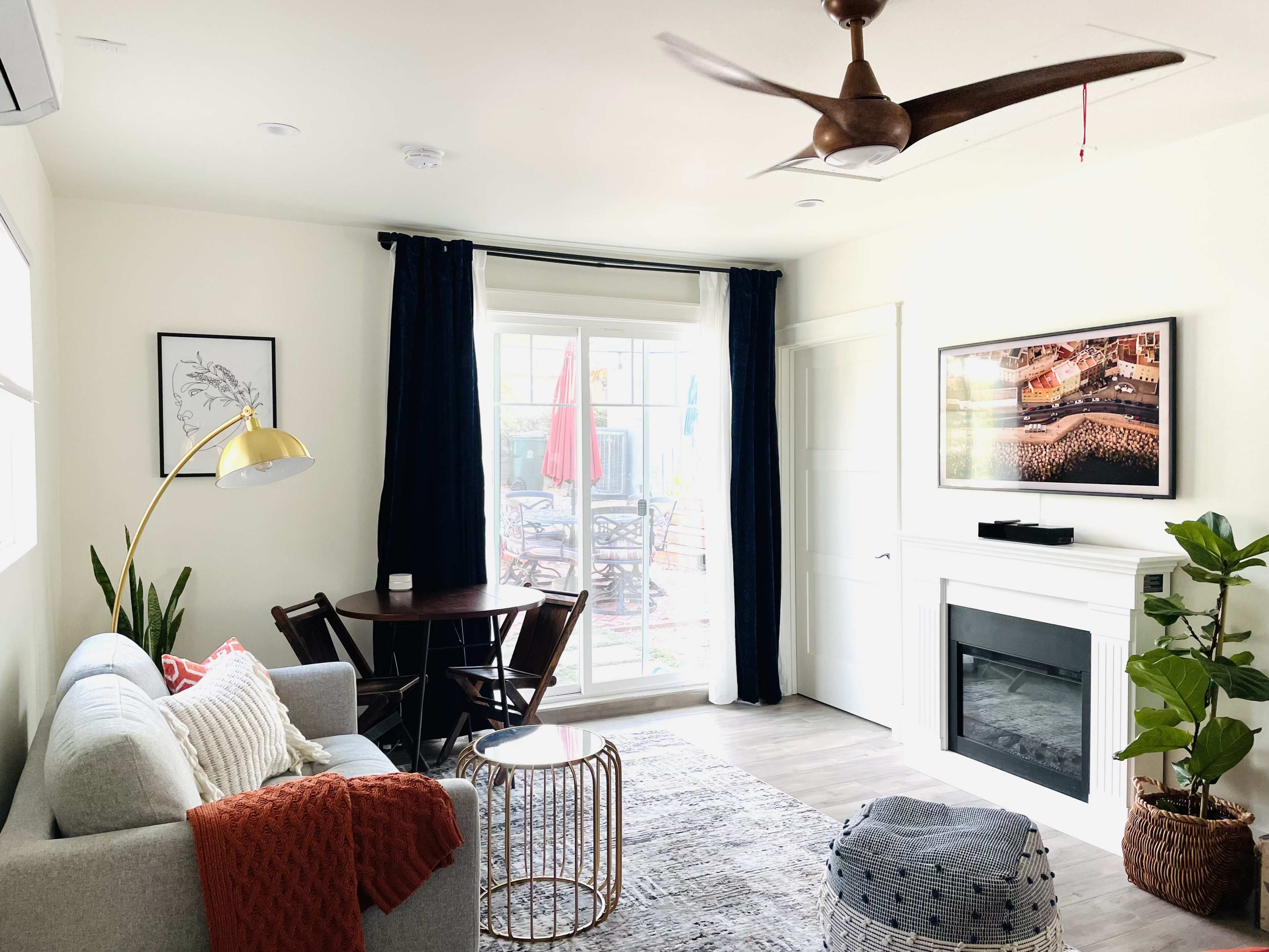 A modern living room features a gray sofa with a red throw, a round coffee table, a dining area, and a view of an outdoor patio through large sliding glass doors.