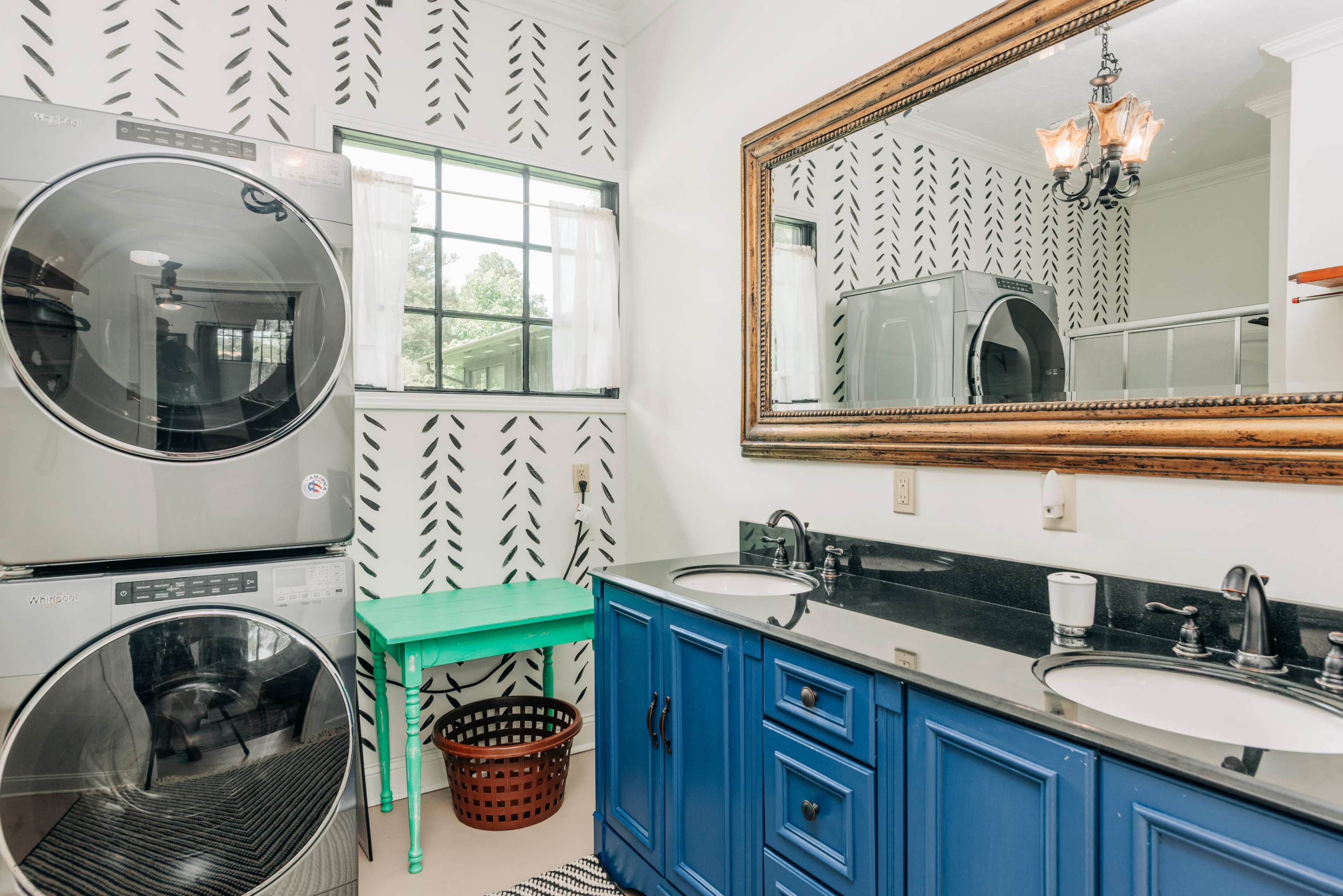 A laundry room with stacked washers and dryers, a blue double vanity with sinks, a green table, and a large mirror against a patterned wall.