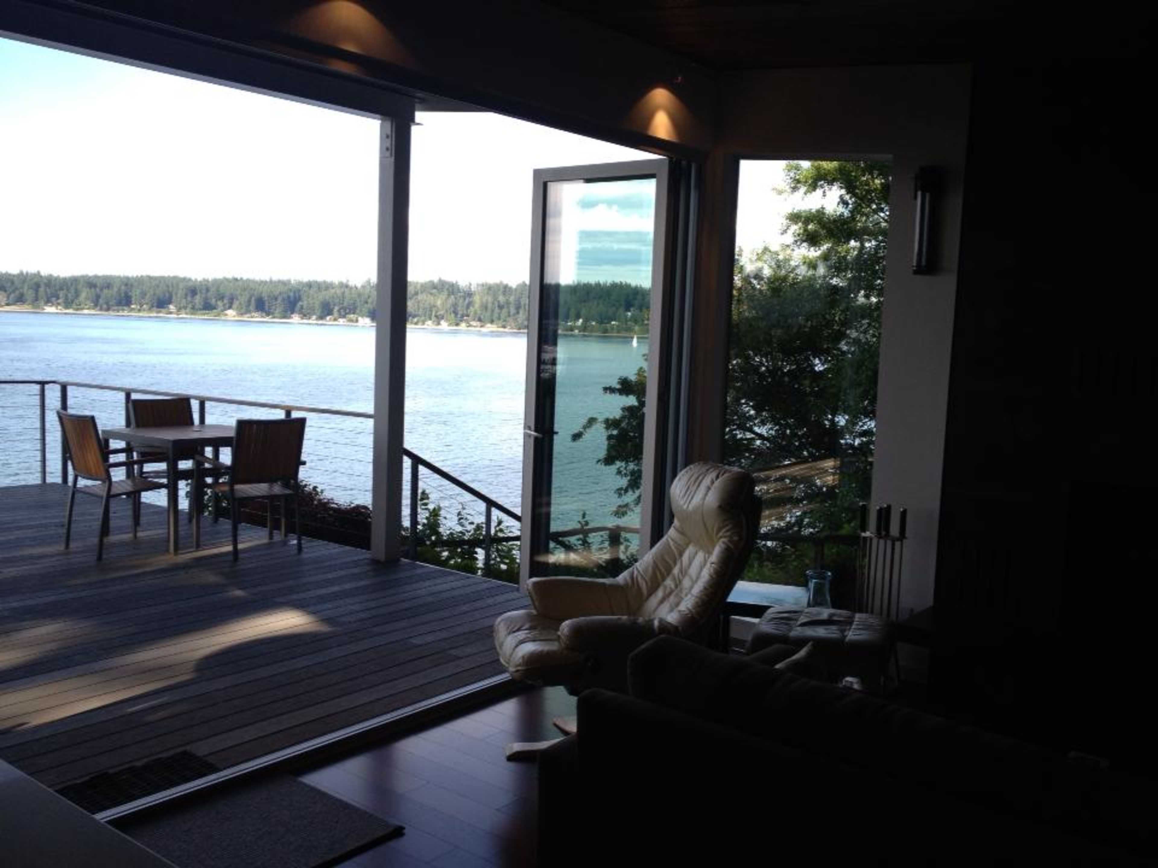 A modern living space opens up to a large deck overlooking a calm body of water with trees in the background.