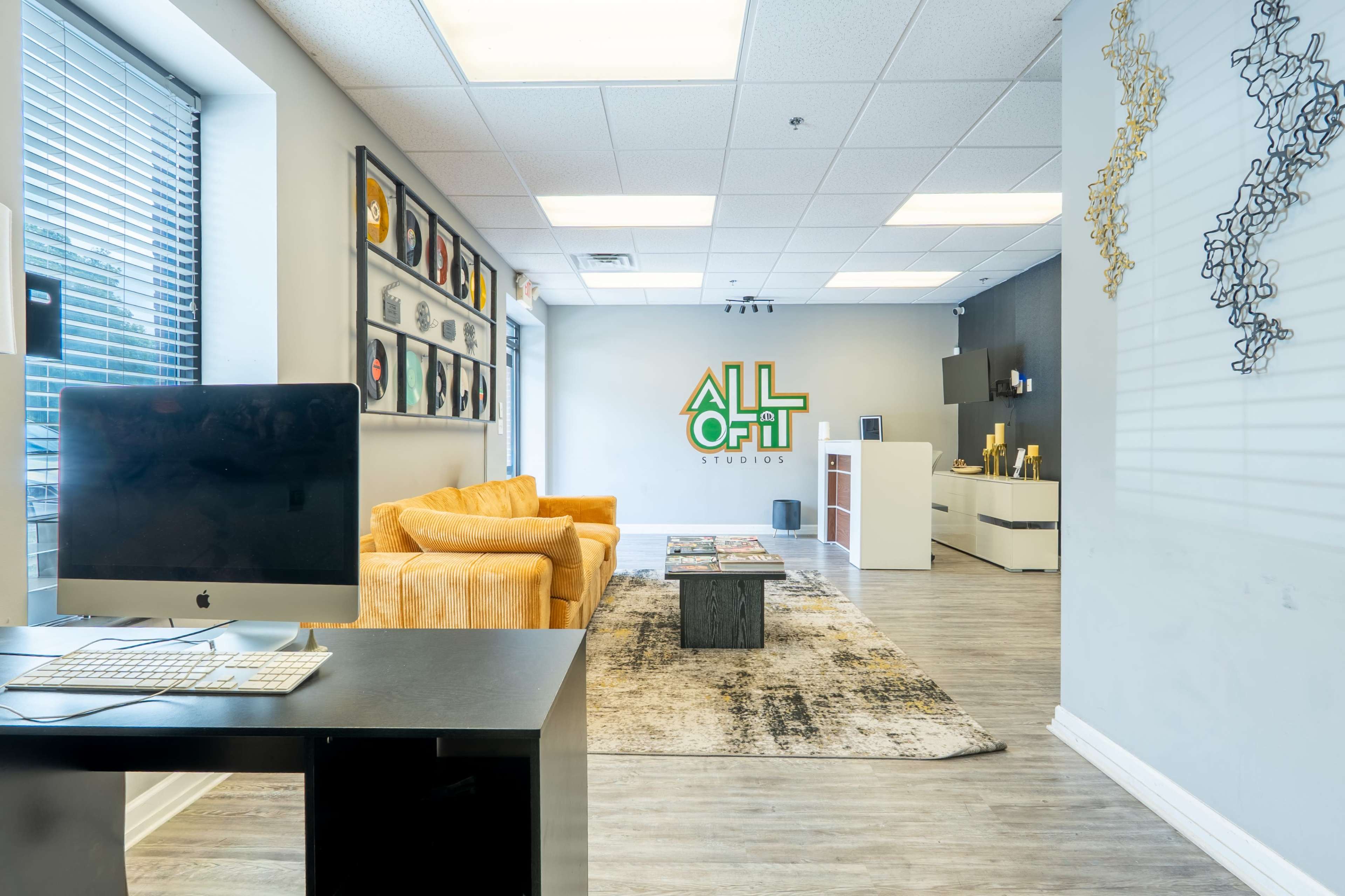 The image shows a modern office space with a reception area featuring a yellow couch, a desk with a computer, framed artwork on the walls, and a prominent sign that reads "ALL OR NOTHING Studios."