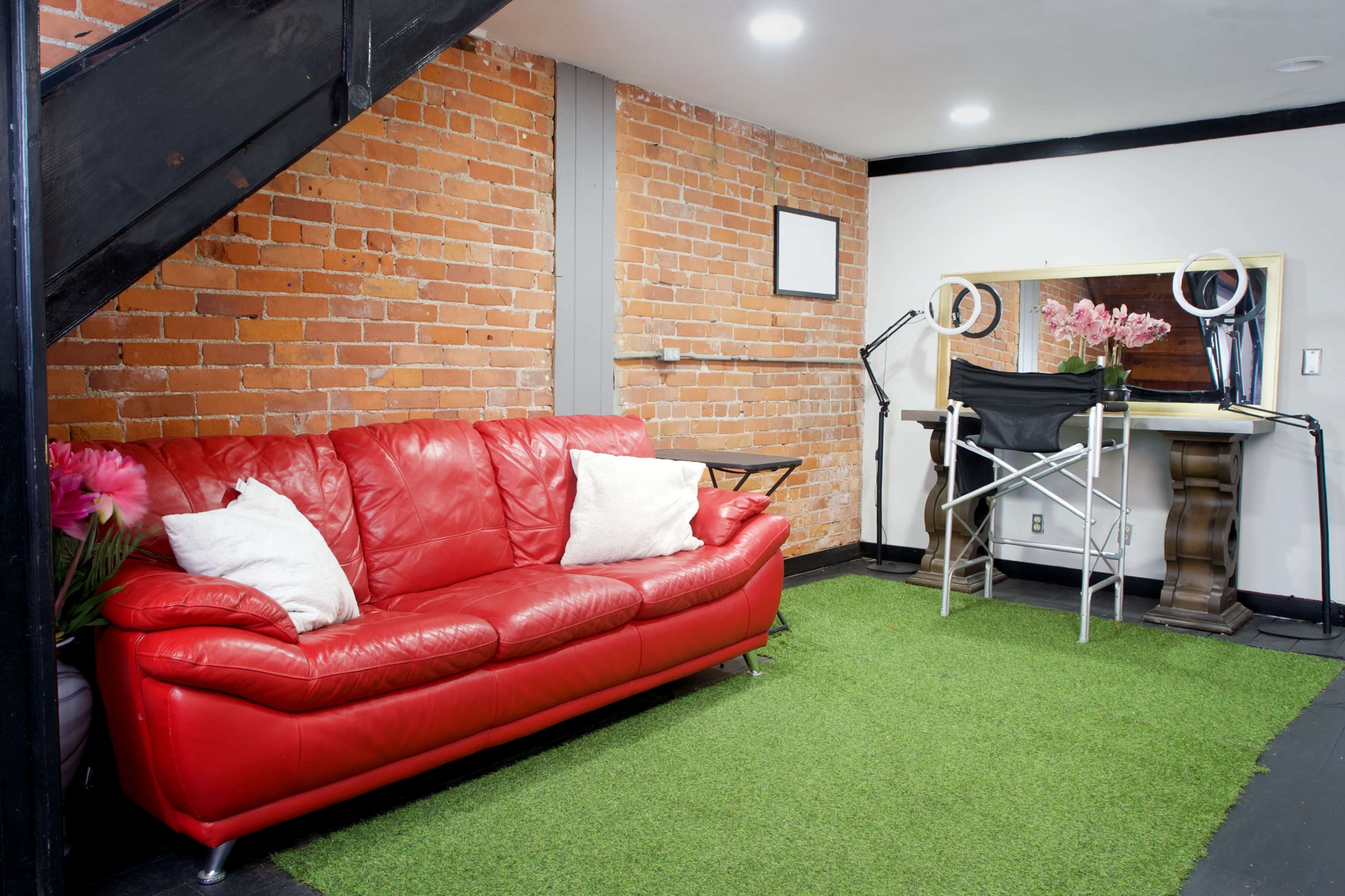 The image shows a cozy interior space with a red leather sofa, artificial grass flooring, and a dressing area featuring a mirror and a table under a staircase.
