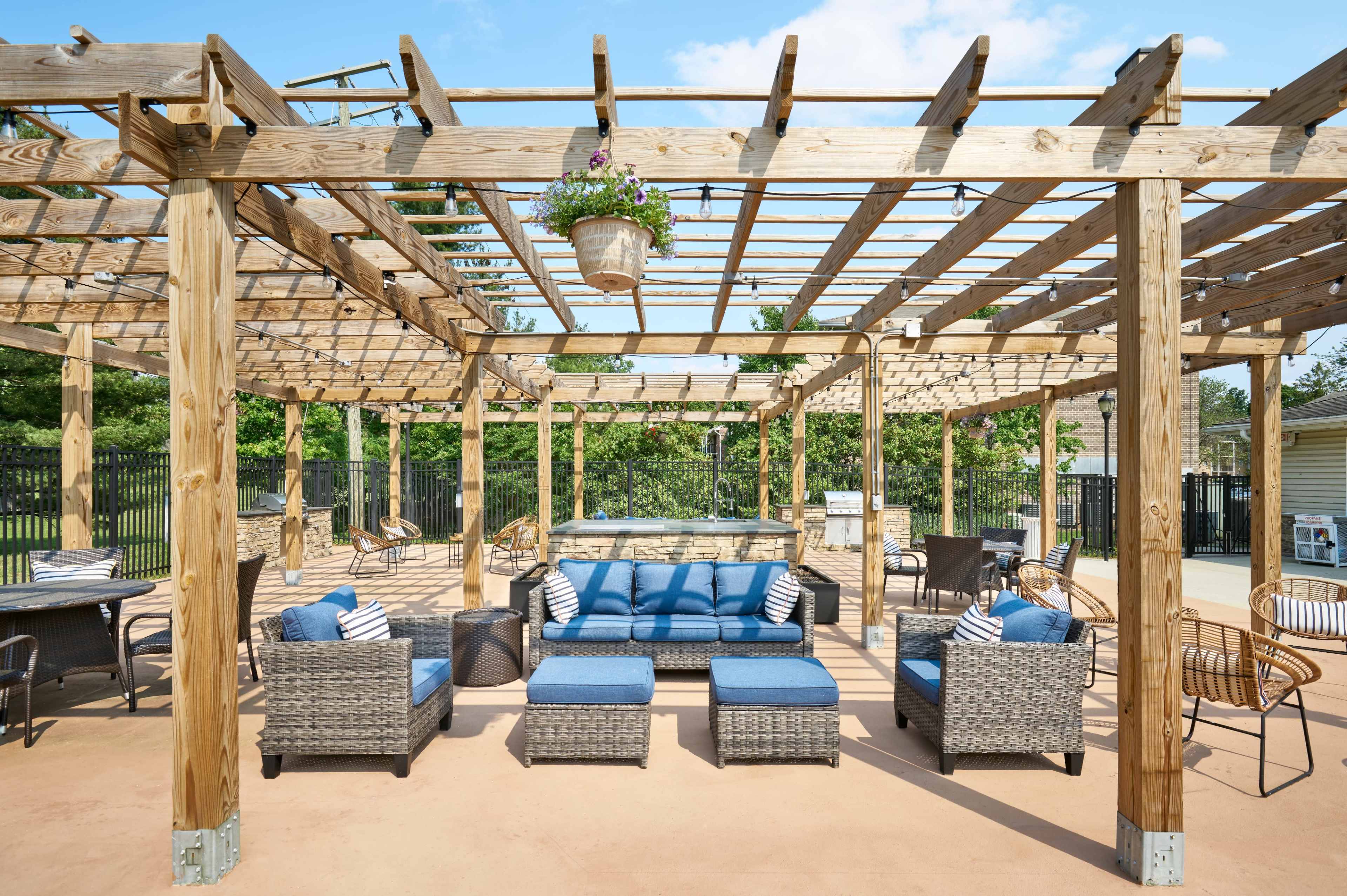 The image shows an outdoor lounge area with a wooden pergola, featuring a seating arrangement with cushions and a table, surrounded by various chairs on a concrete floor.