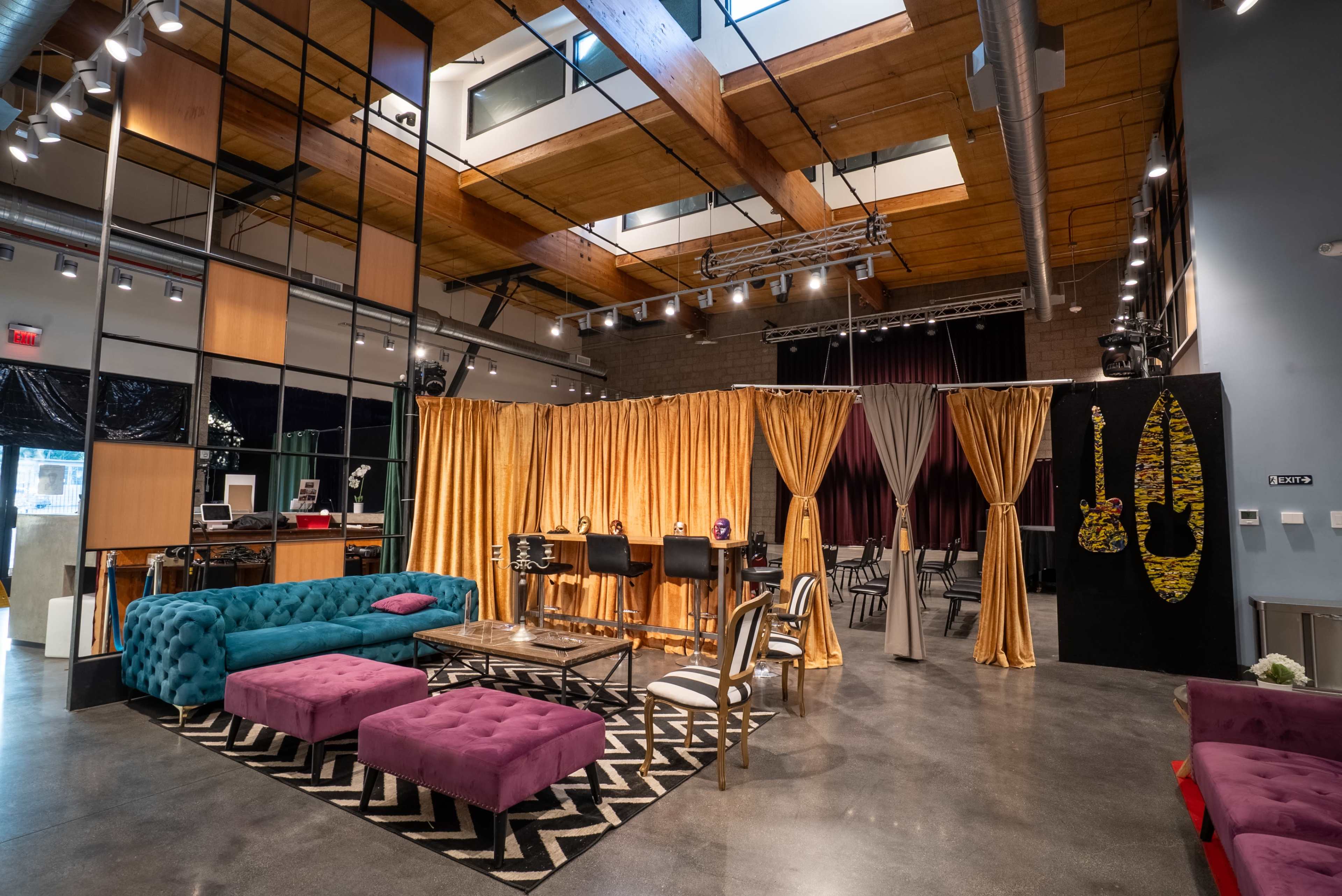 The image shows a modern interior space featuring a seating area with colorful sofas, a performance stage with curtains, and decorative accents on the walls.