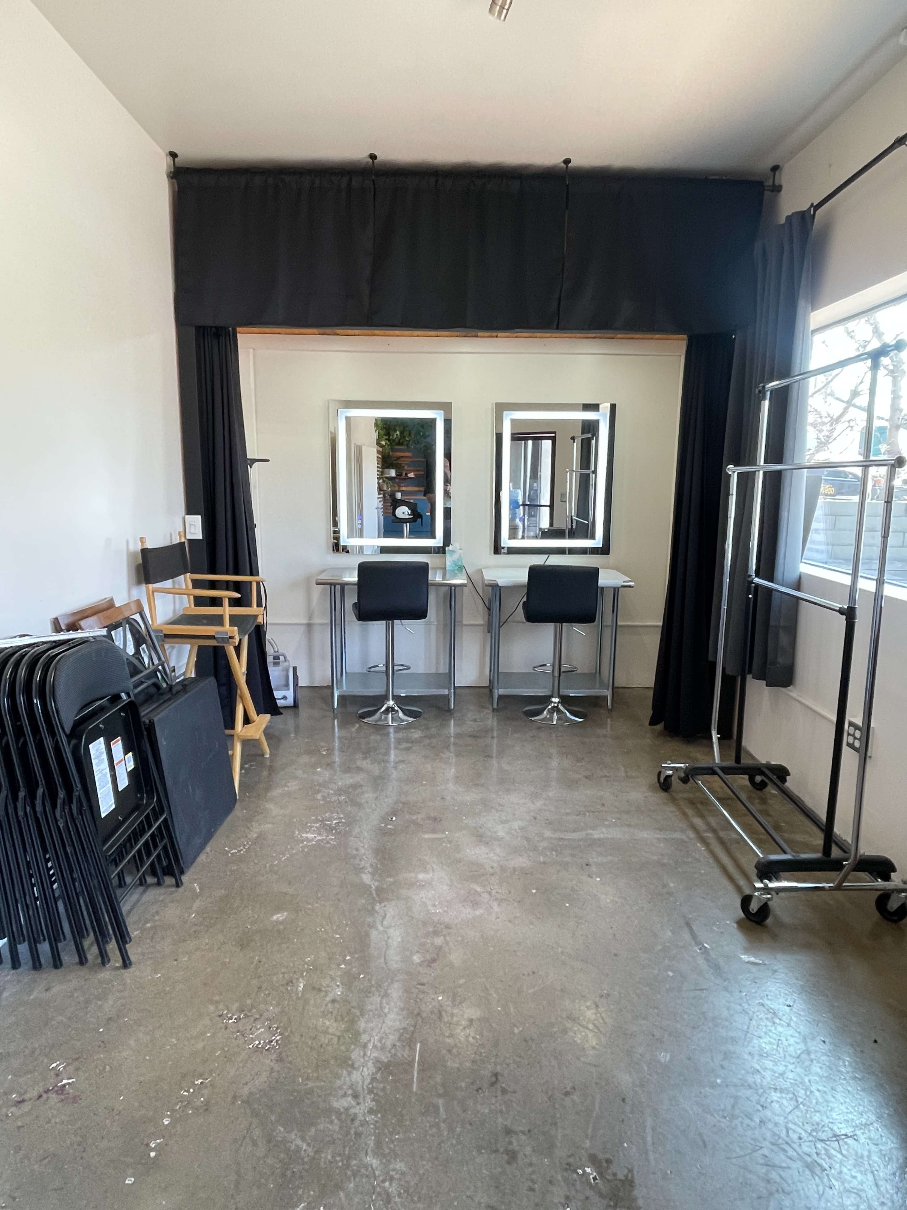 The image shows a well-lit room with two mirrors on a table, side chairs, and a clothing rack, along with several folded chairs stacked in the corner.