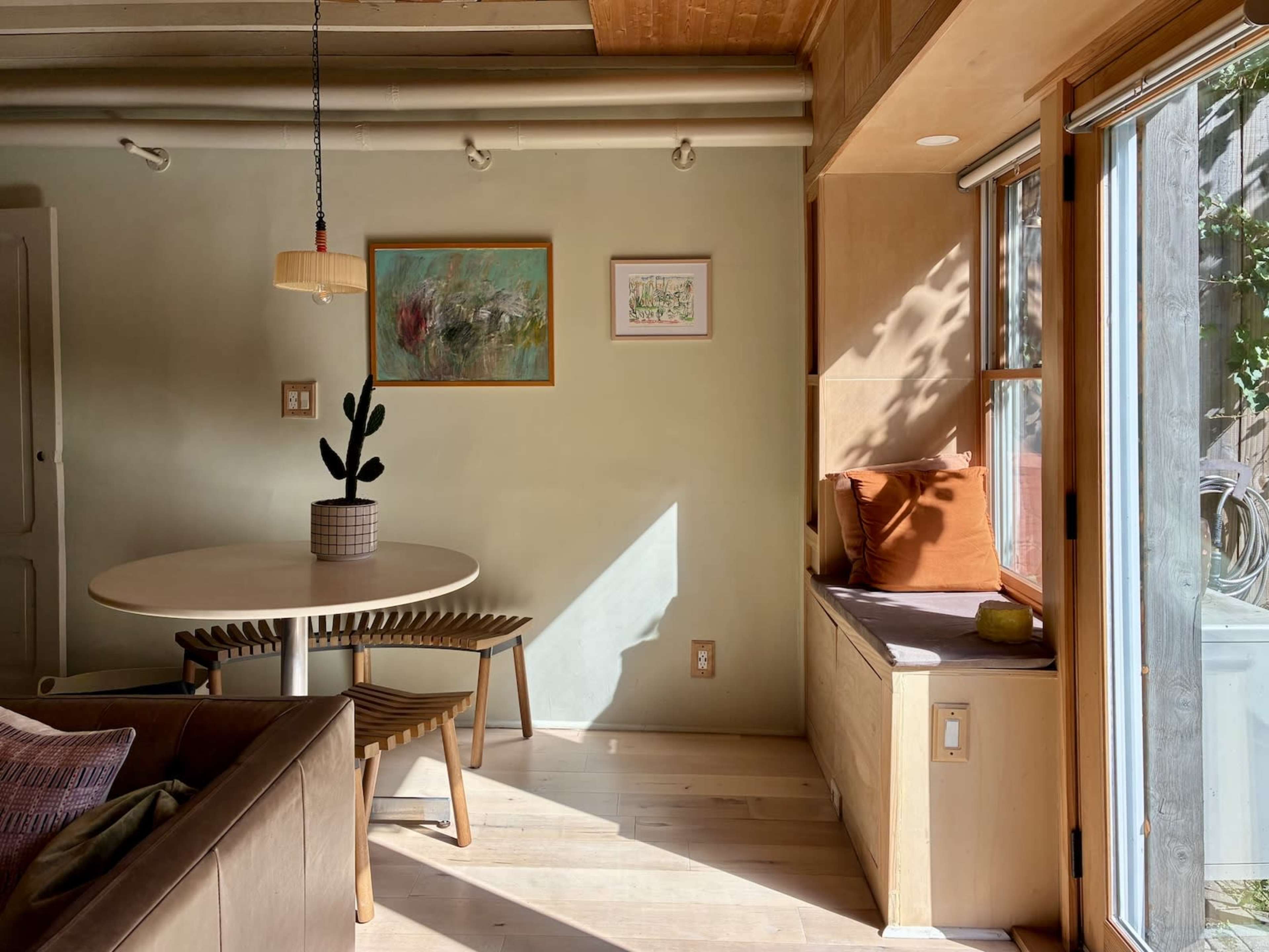 A well-lit modern living space features a round table with wooden chairs, a comfortable sofa, and a window seat filled with cushions.