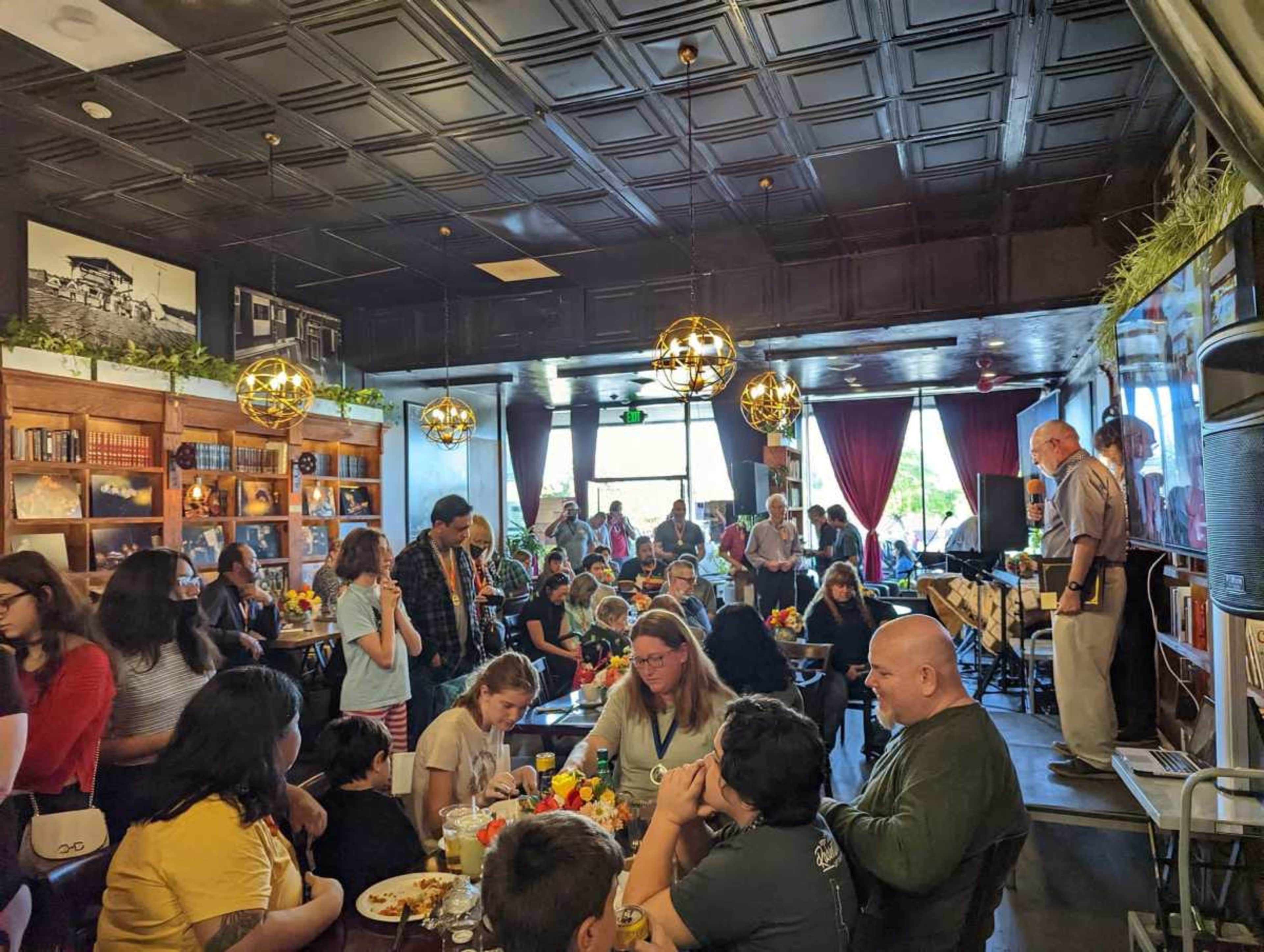 A crowded café features tables filled with patrons while a small stage displays a musician performing in the background.