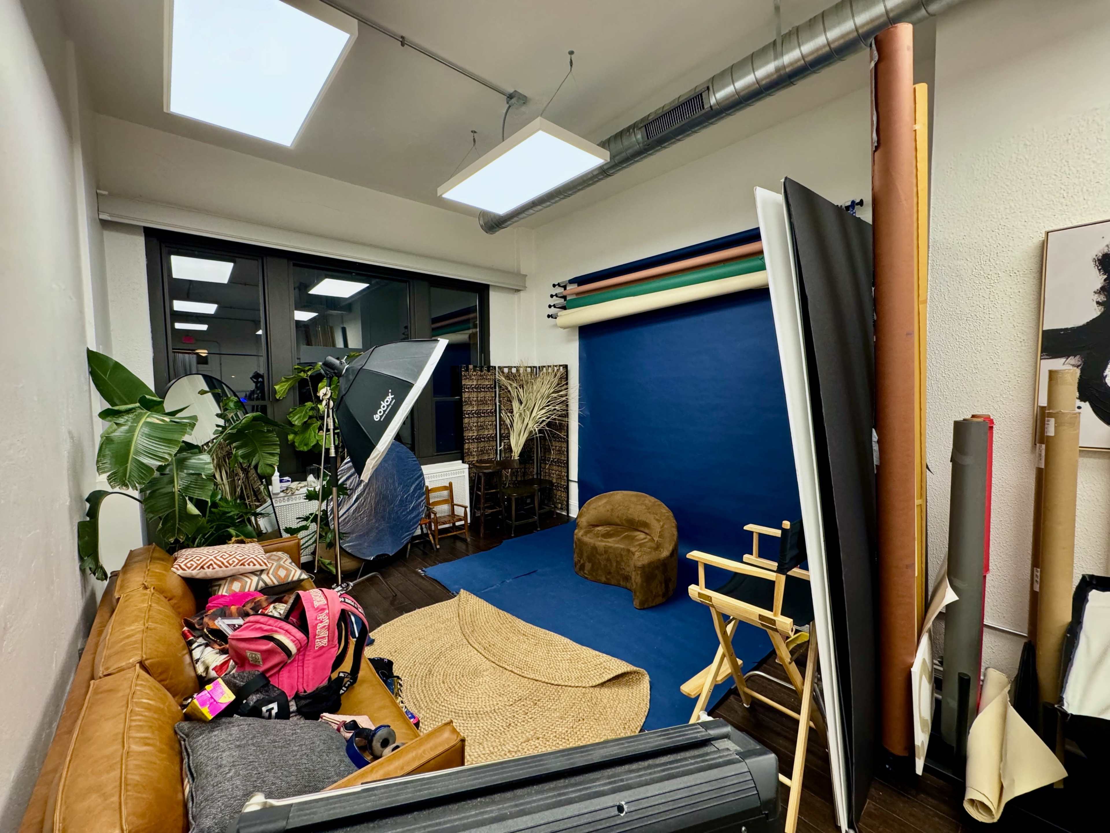 A photography studio featuring a blue backdrop, assorted props, a large umbrella light, and a brown chair, with various items scattered on a couch.