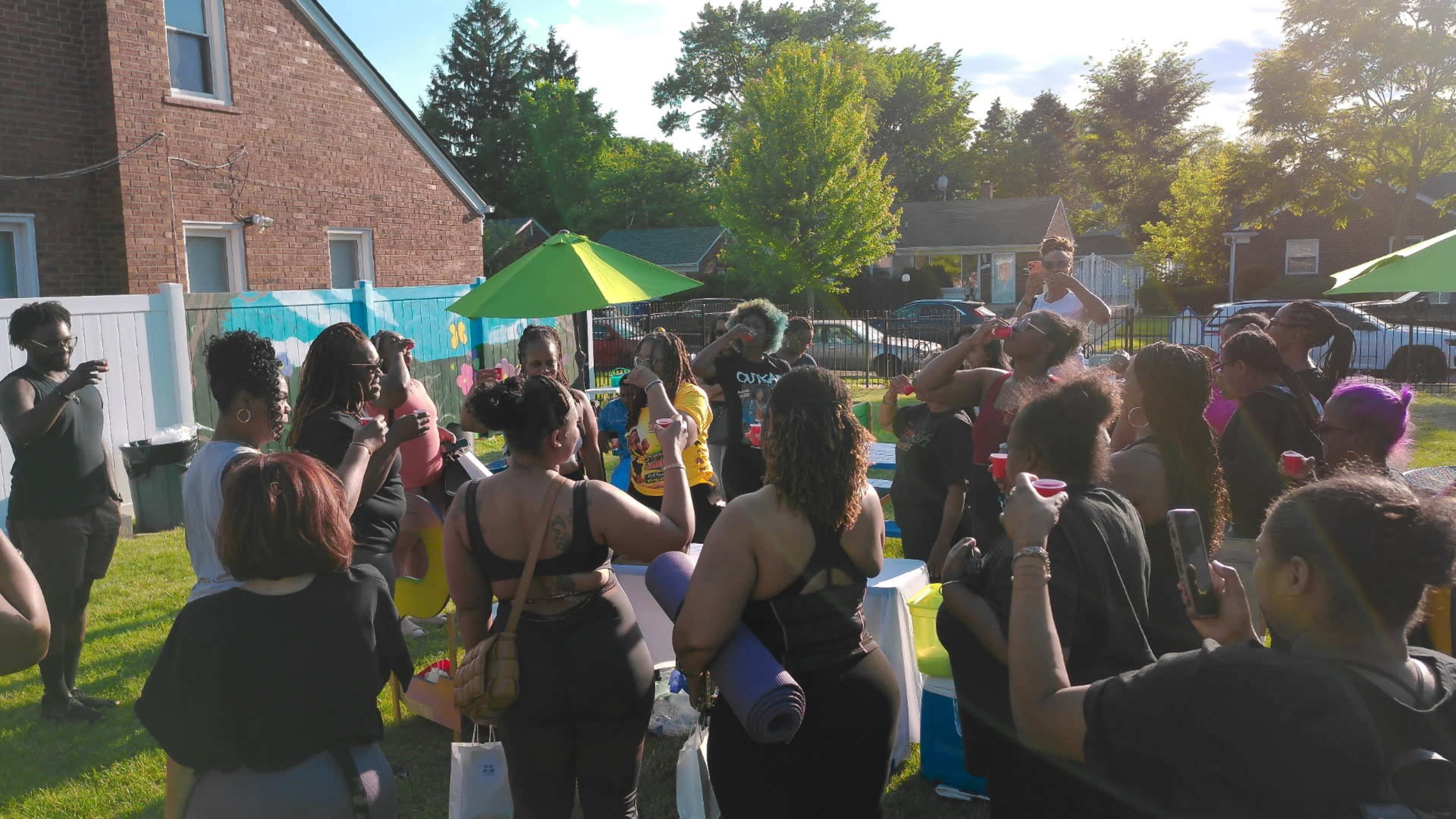 A group of individuals gathers outdoors, participating in an event with colorful decorations under green umbrellas.