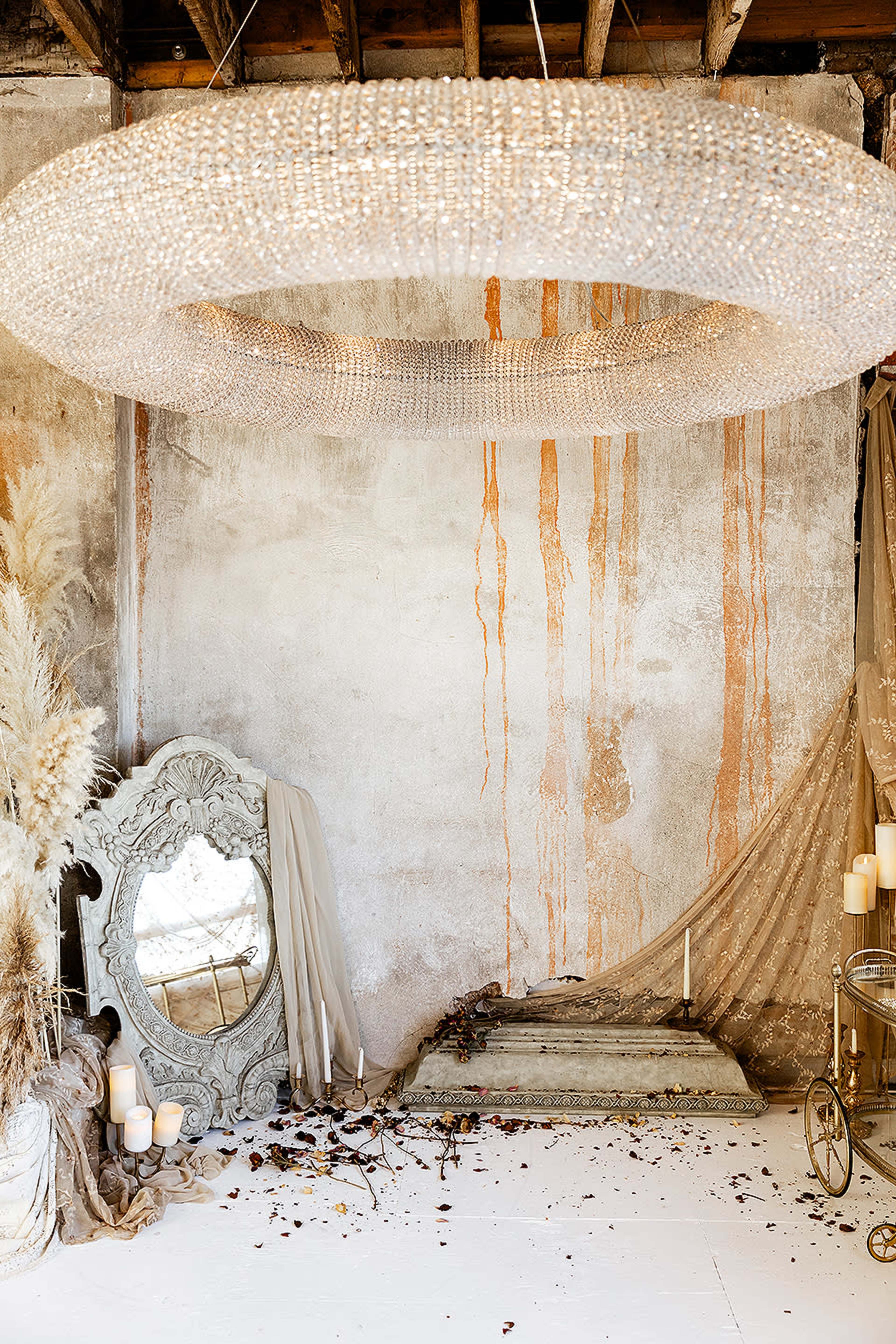 A large circular chandelier hanging above a room with a distressed wall, a vintage mirror, scattered candles, and draped fabric on the floor.