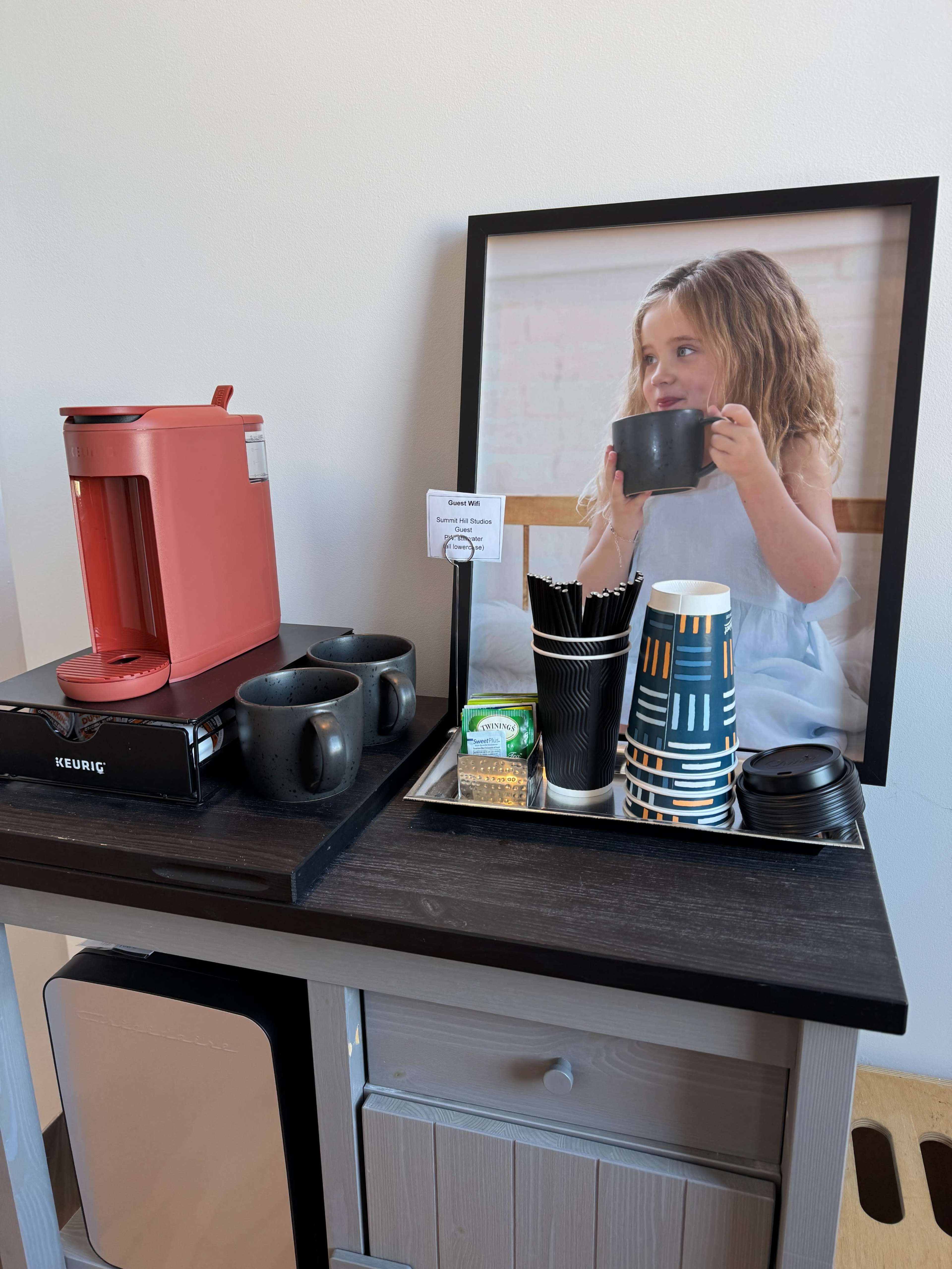 The image shows a coffee station featuring a red Keurig machine, cups, stirrers, and a small tray with a snack, alongside a framed picture of a girl holding a cup.