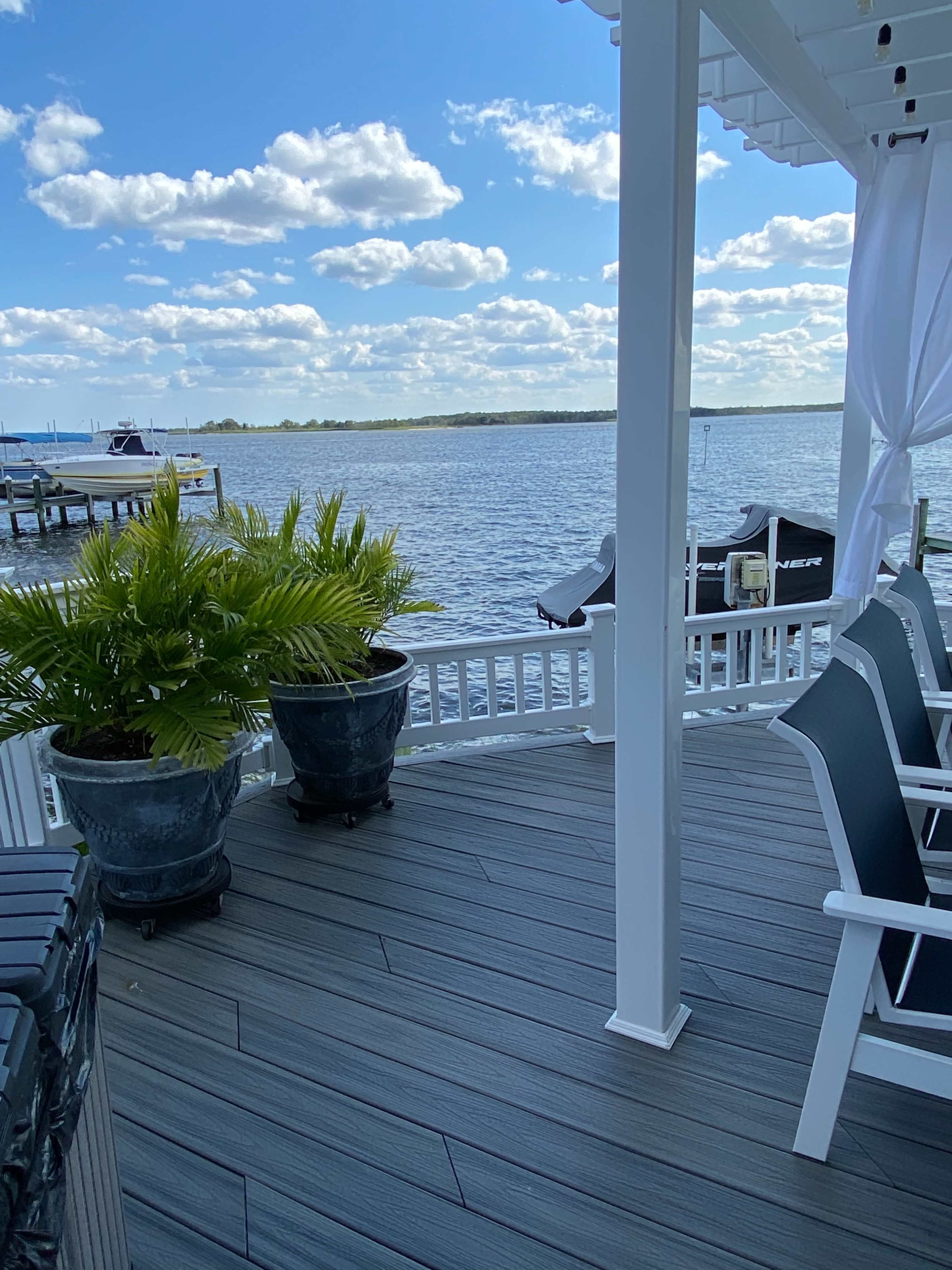 The image shows a riverfront deck area with two potted ferns and black chairs overlooking the water.