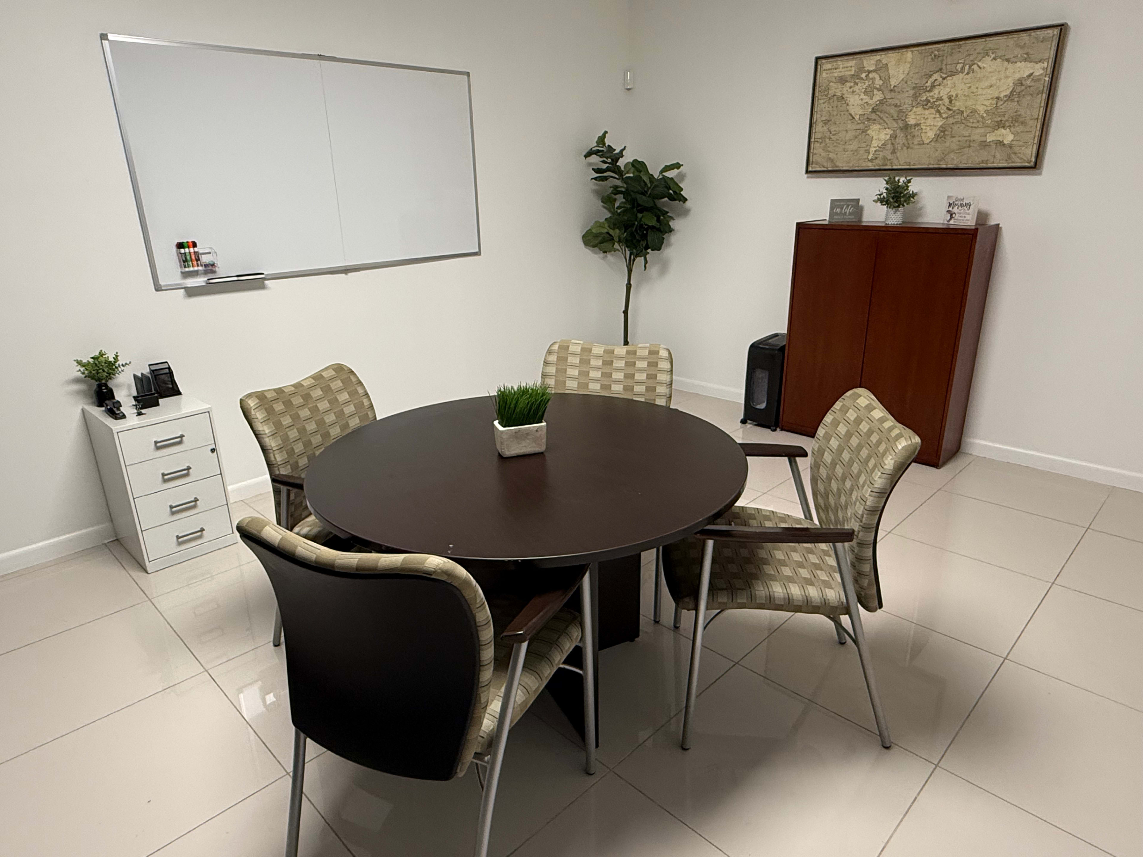 A round table surrounded by four patterned chairs is set in a small meeting room with a whiteboard, a plant, and a cabinet against the wall.