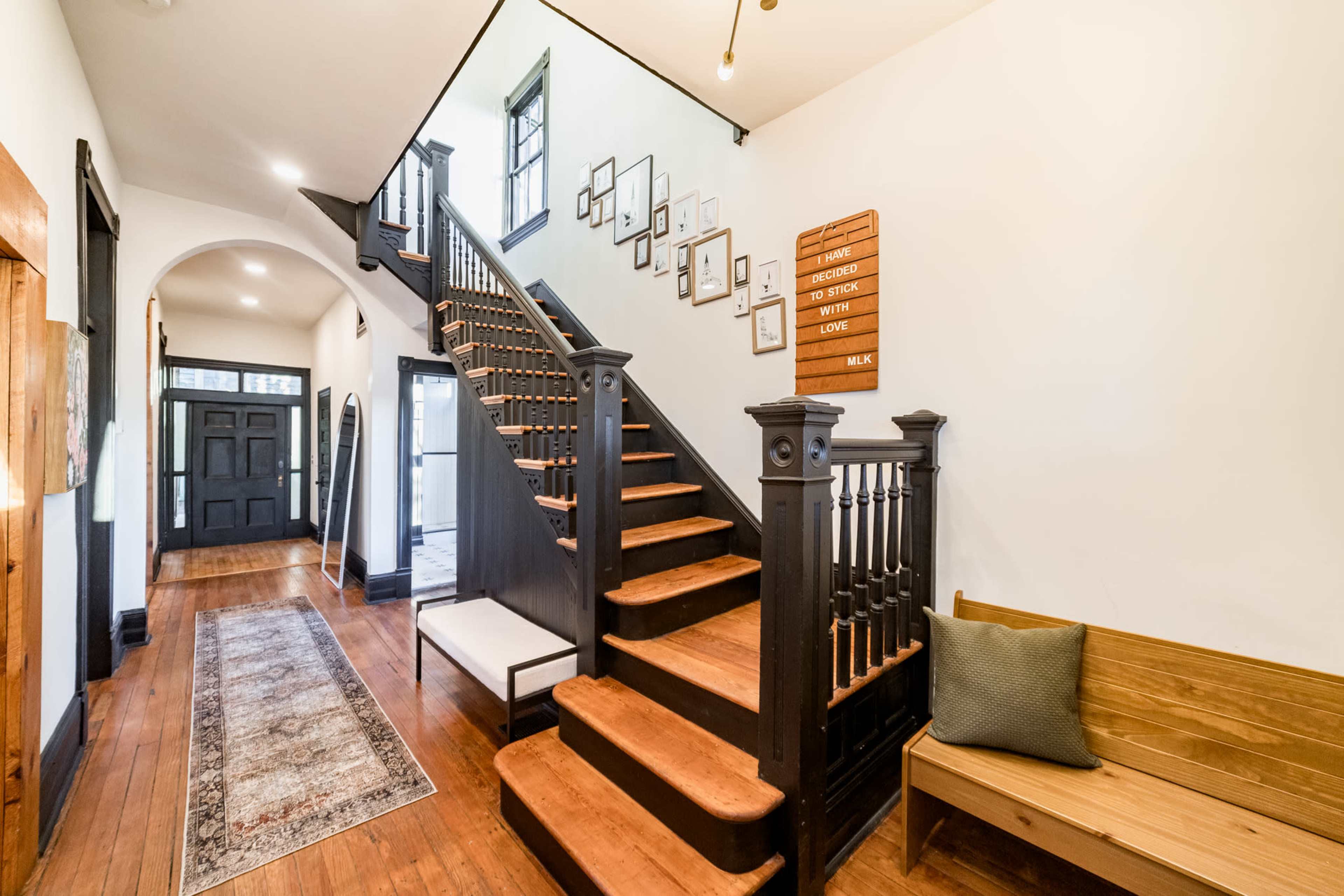 A well-lit hallway featuring a staircase with wooden steps, black banisters, framed pictures on the wall, and a wooden bench.