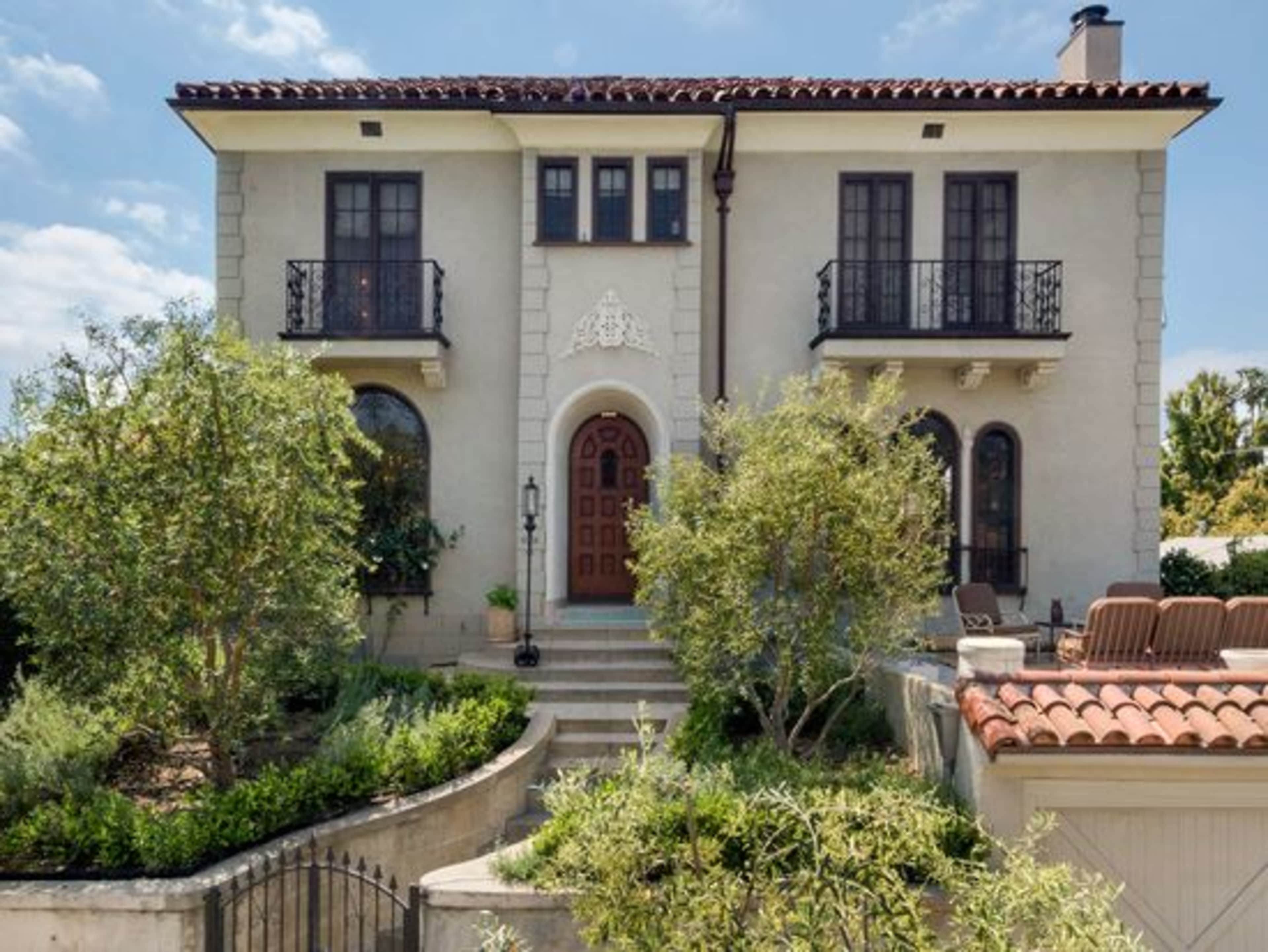 A two-story Mediterranean-style house features a central wooden door, balconies on the upper level, and landscaped greenery in the front yard.