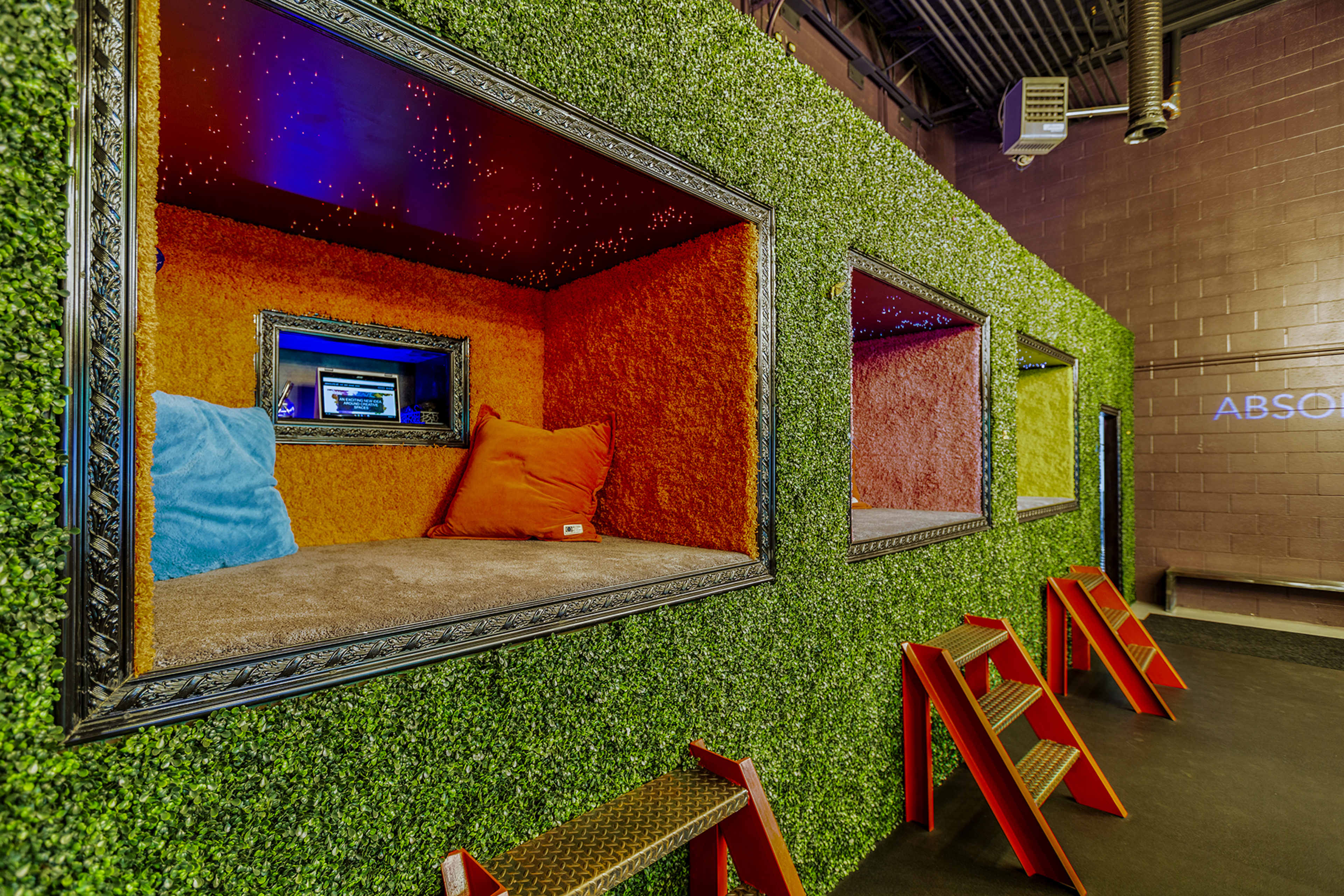 Colorful, framed lounging areas built into a green, grass-like wall, featuring bright pillows and a cozy aesthetic.