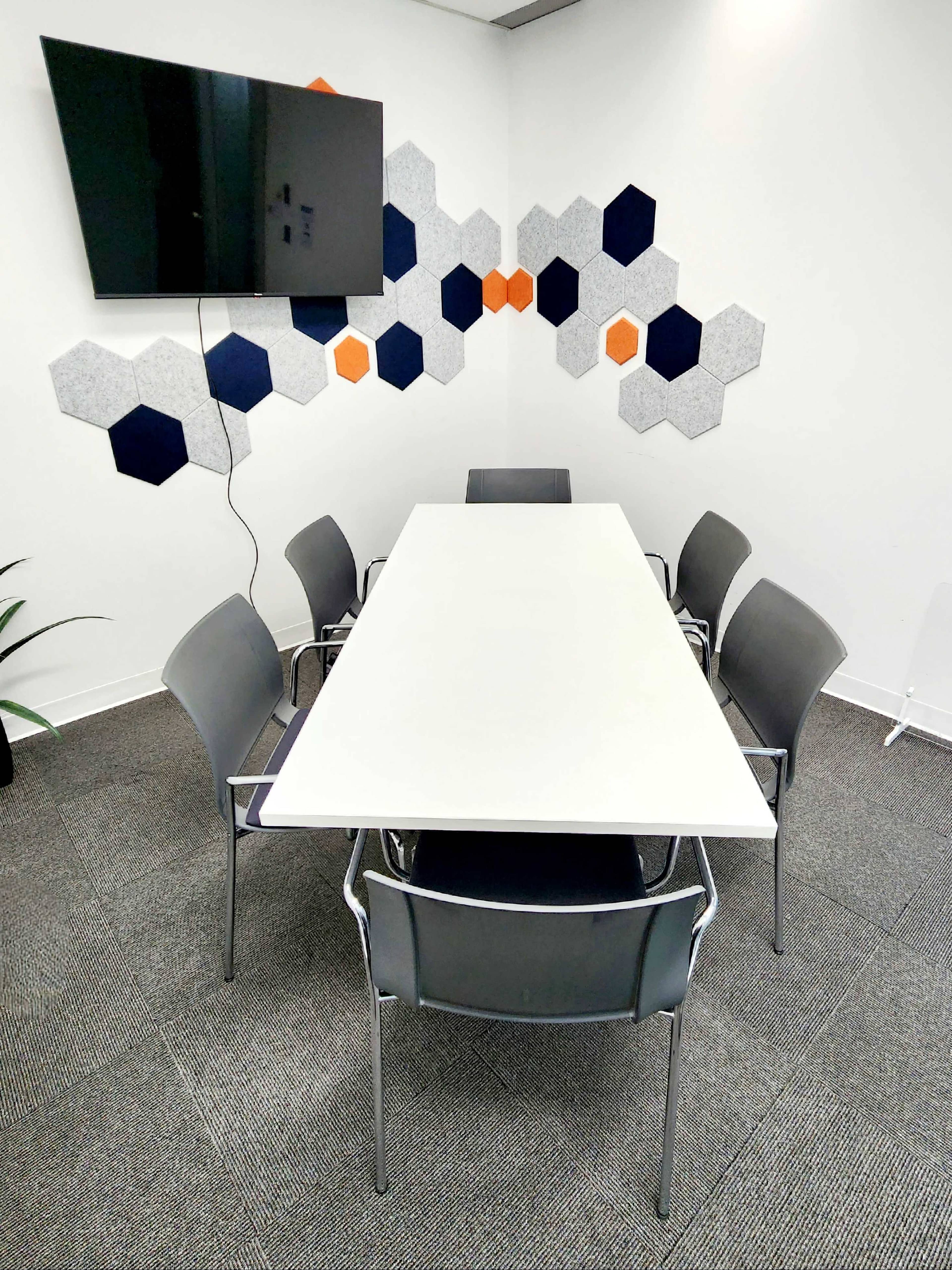 A conference room features a white table surrounded by six gray chairs, with a television mounted on the wall and hexagonal wall decor in shades of navy and orange.