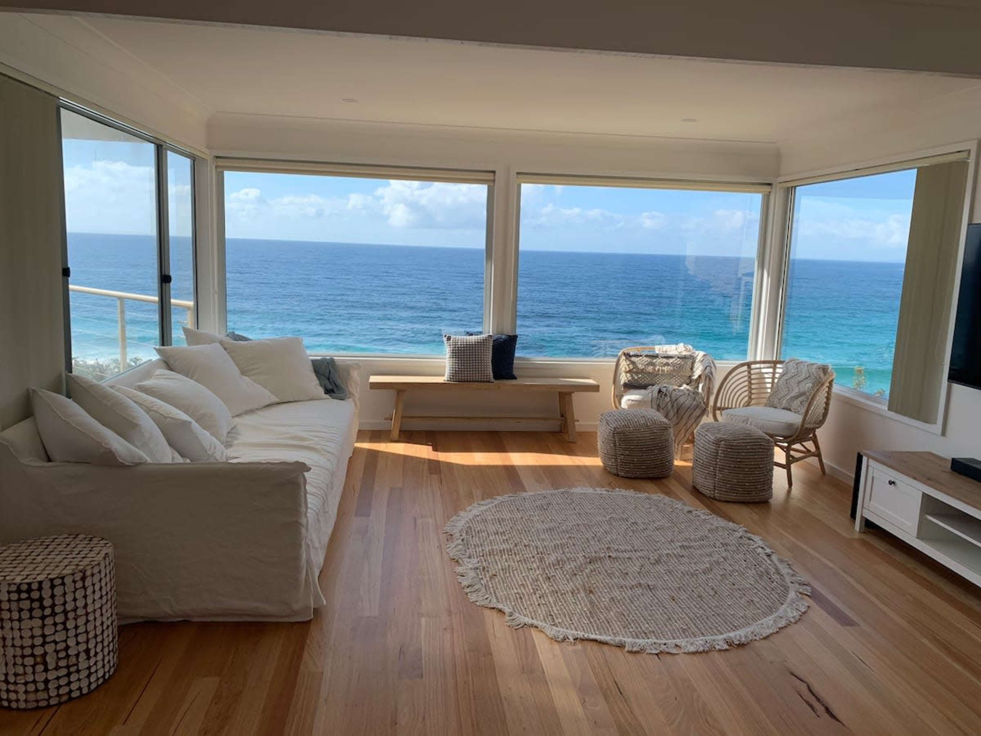 A bright living room with large windows overlooking the ocean, featuring a white sofa, a wooden bench, and woven chairs on a wooden floor.