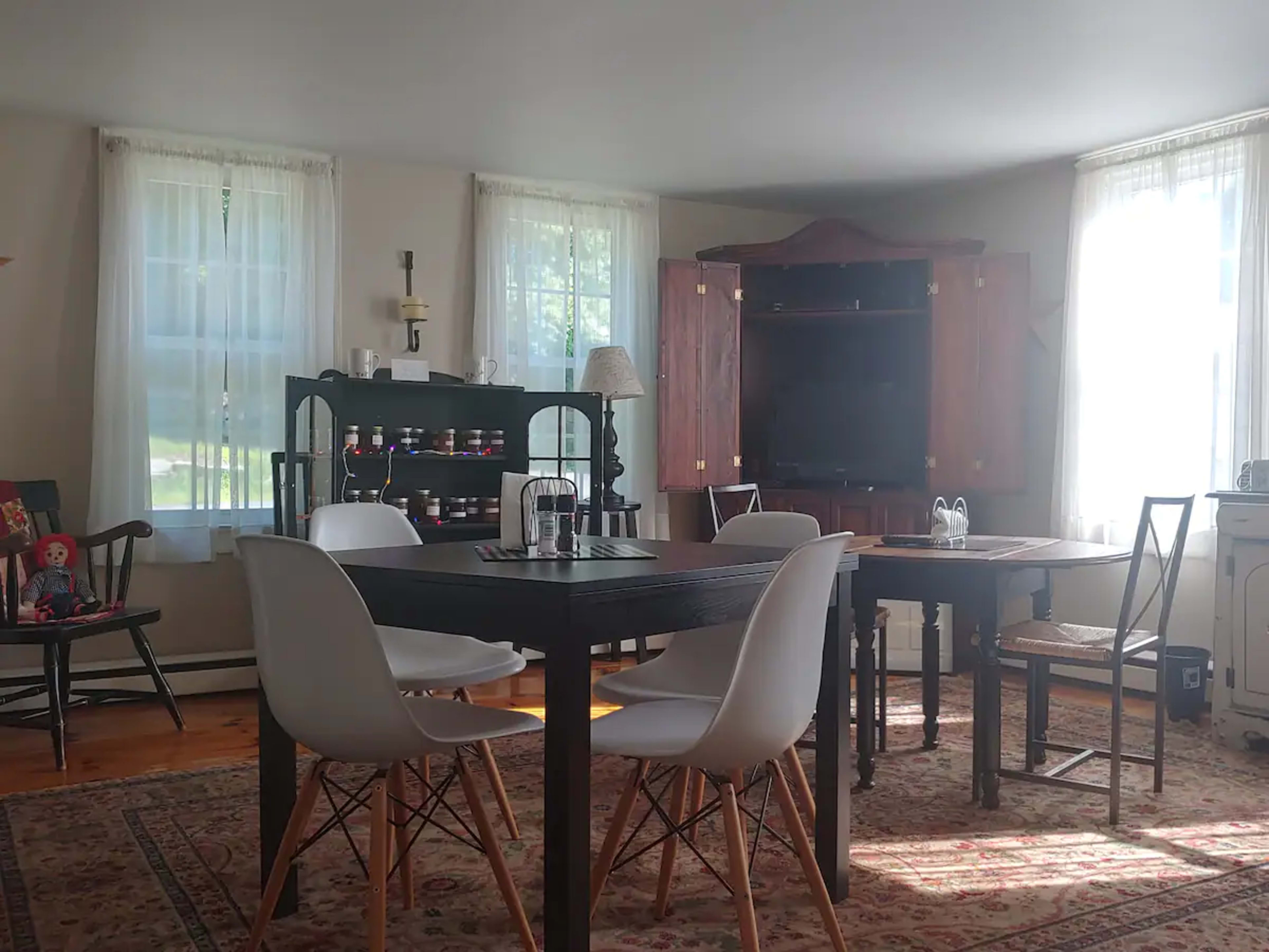 A bright, inviting room with a wooden dining table surrounded by modern chairs, a rocking chair, a cabinet with television, and several windows allowing natural light to fill the space.