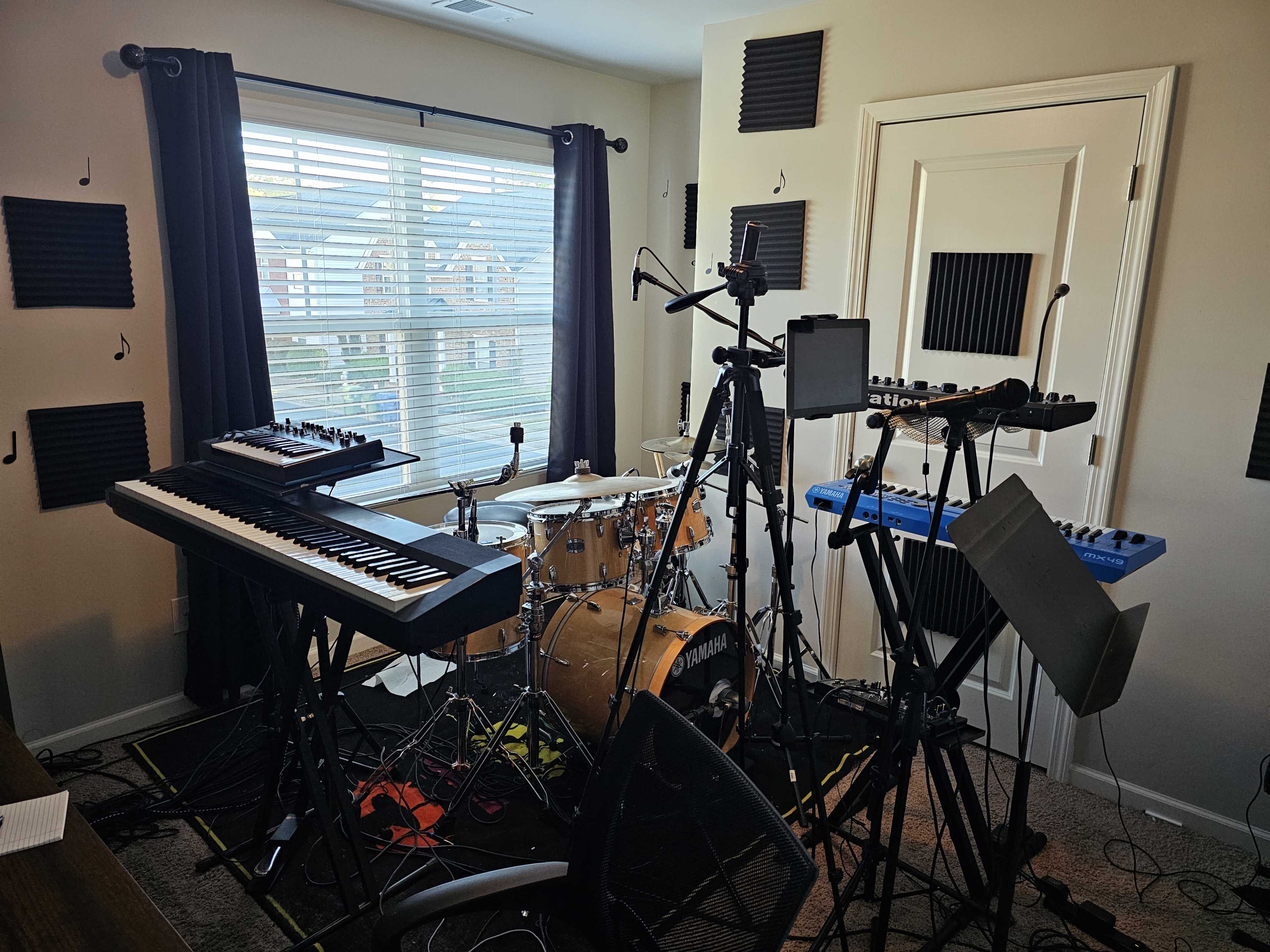 A music studio filled with various musical instruments, including keyboards, a drum set, and audio equipment, in a room with a window and soundproofing panels on the walls.