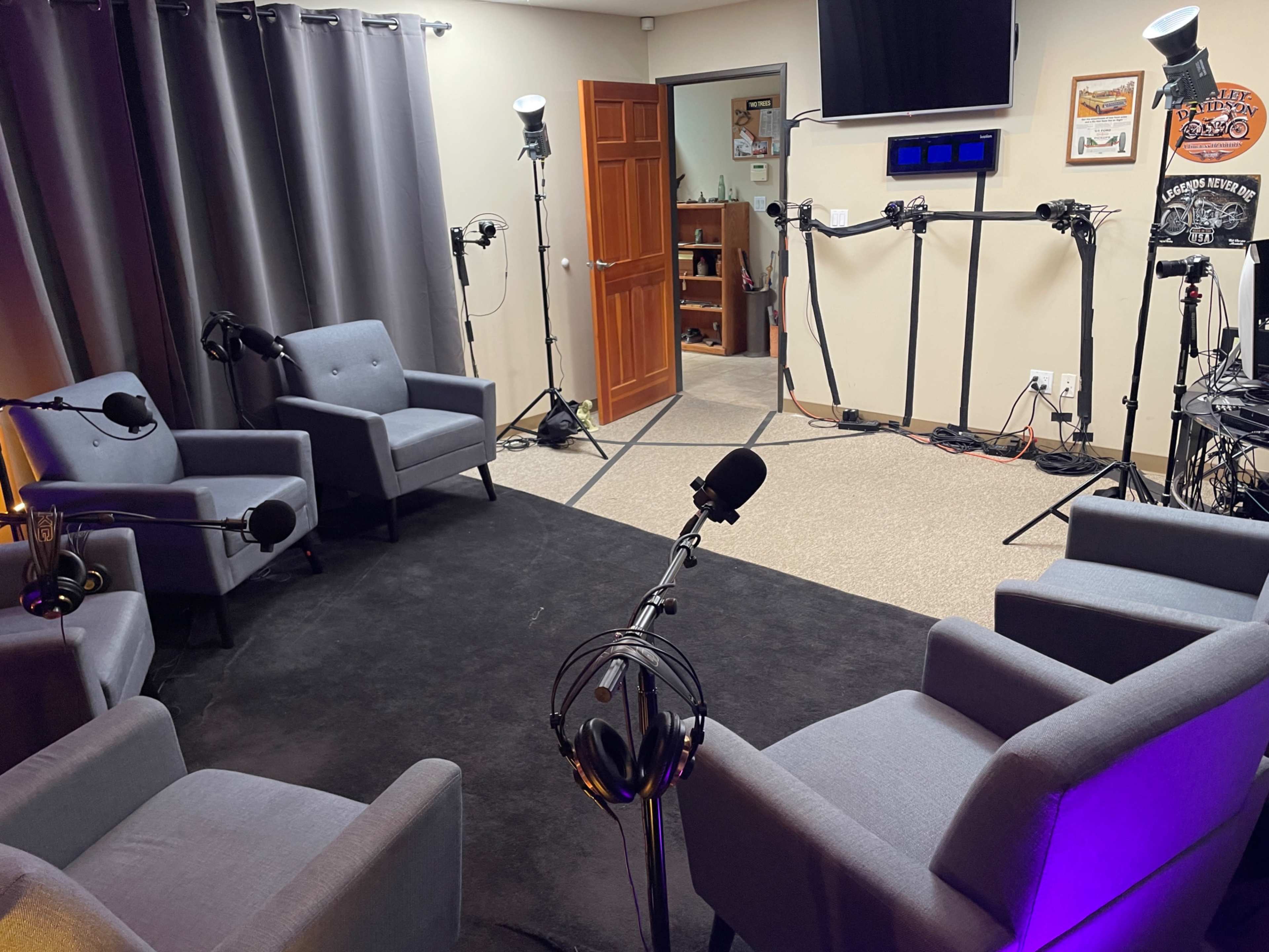 The image shows a recording studio with four gray chairs arranged in a circle, surrounded by microphones on stands and soft lighting.