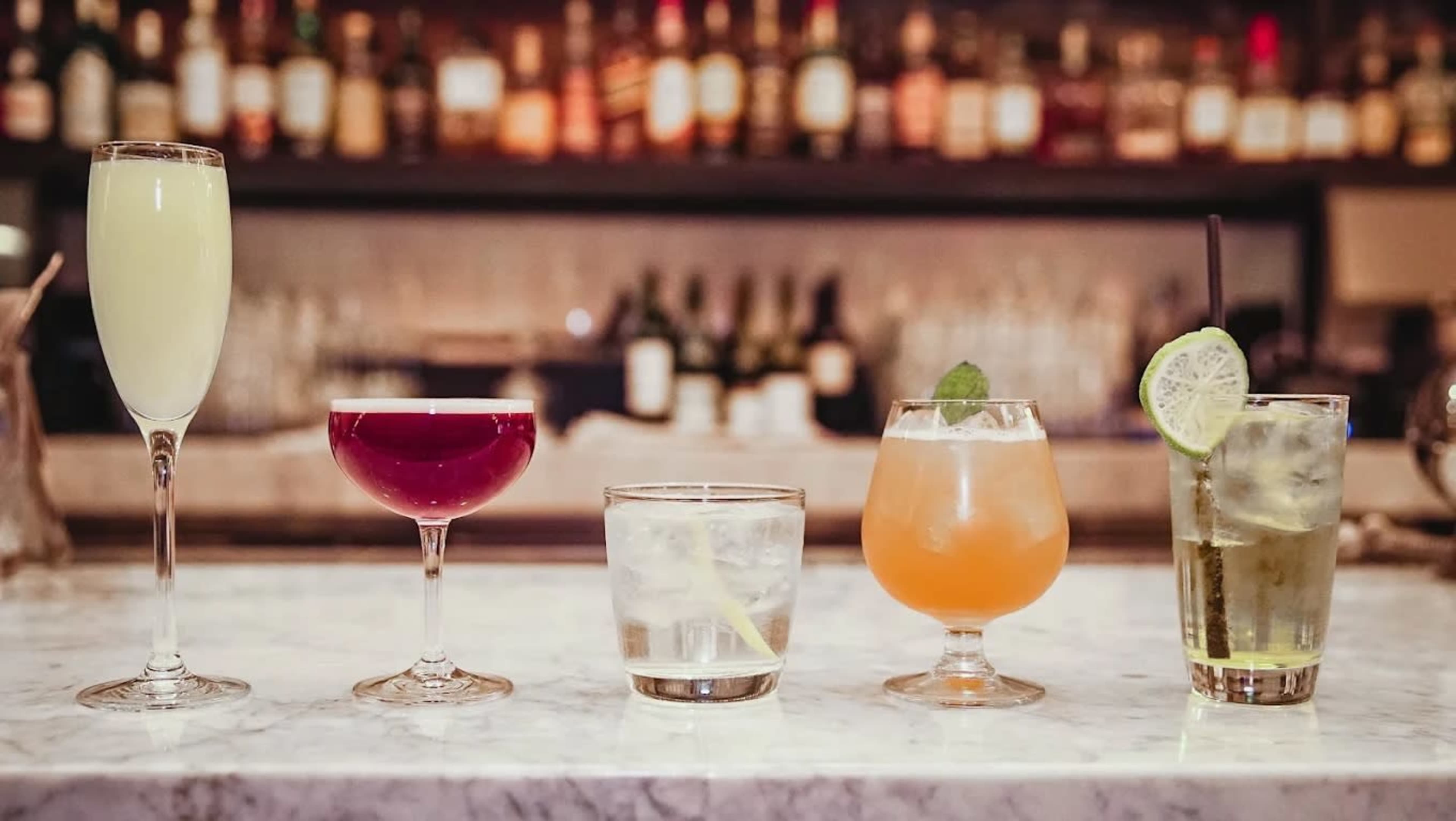 A lineup of five different cocktails is displayed on a marble bar counter with various bottles of liquor in the background.