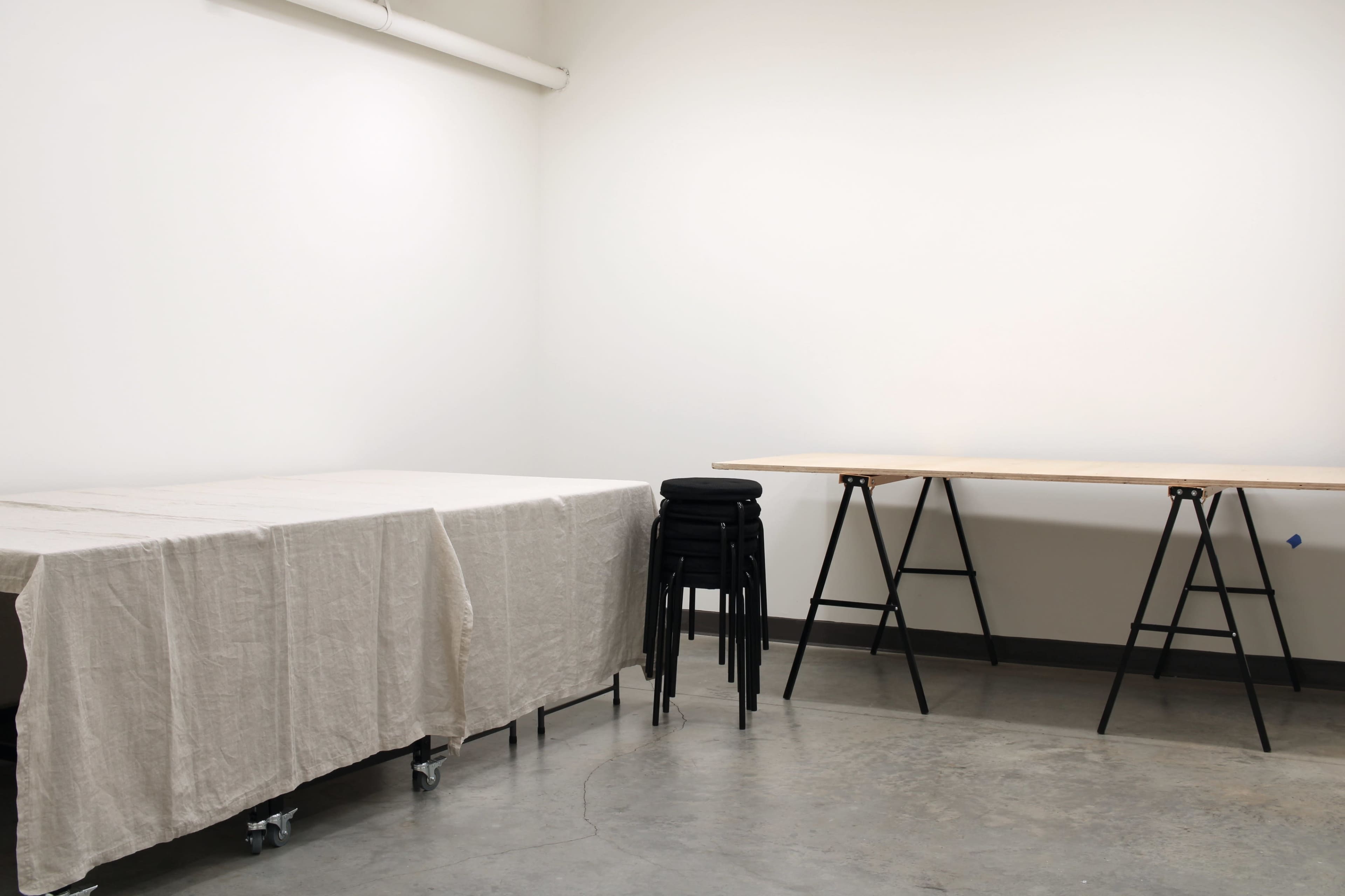 A sparsely furnished room with a table covered in a beige cloth and a stack of black chairs near a wooden table on sawhorse legs.