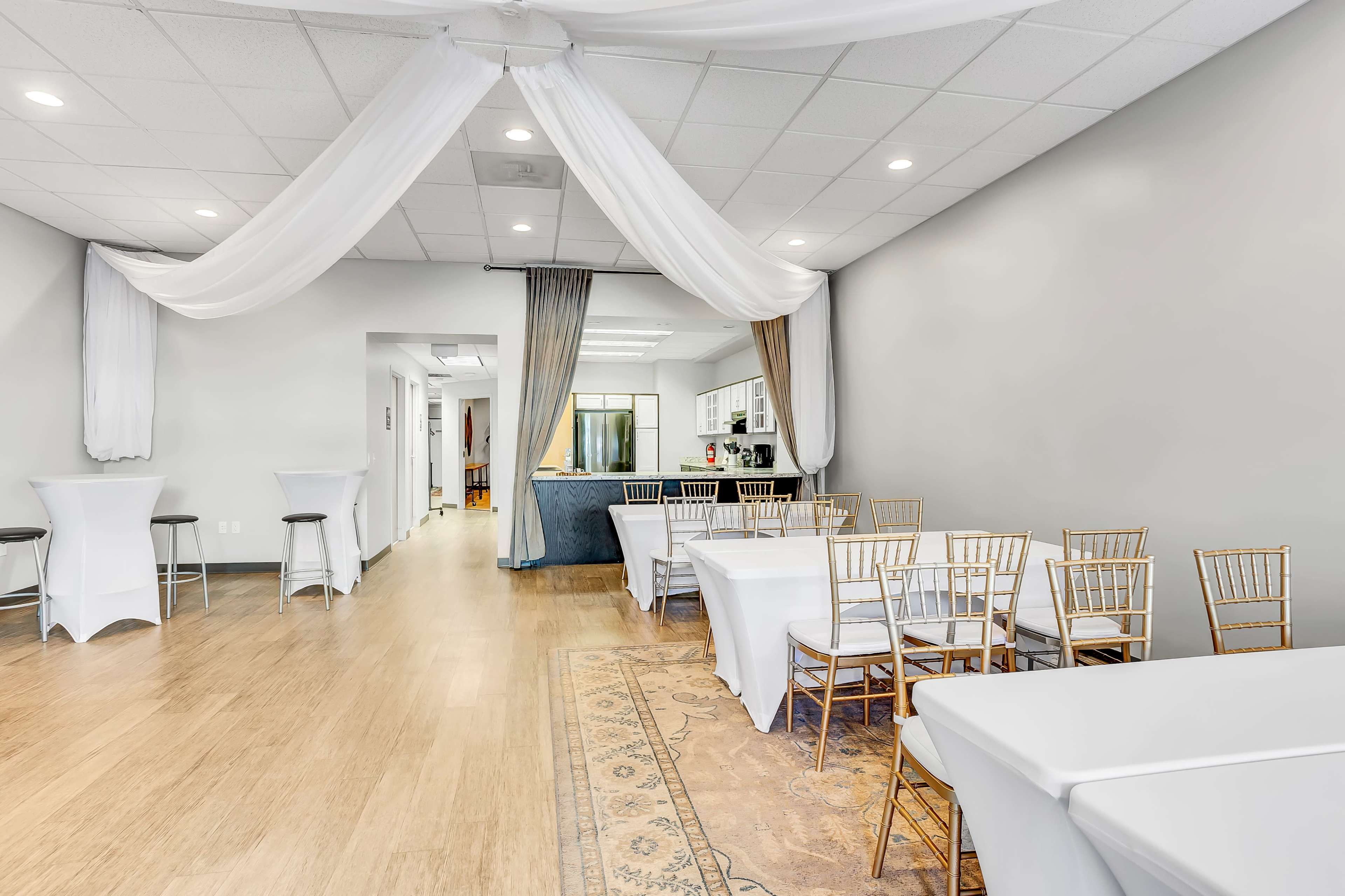 A spacious event room with white tables and chairs arranged for seating, accented by draped fabric overhead and a kitchen area visible in the background.