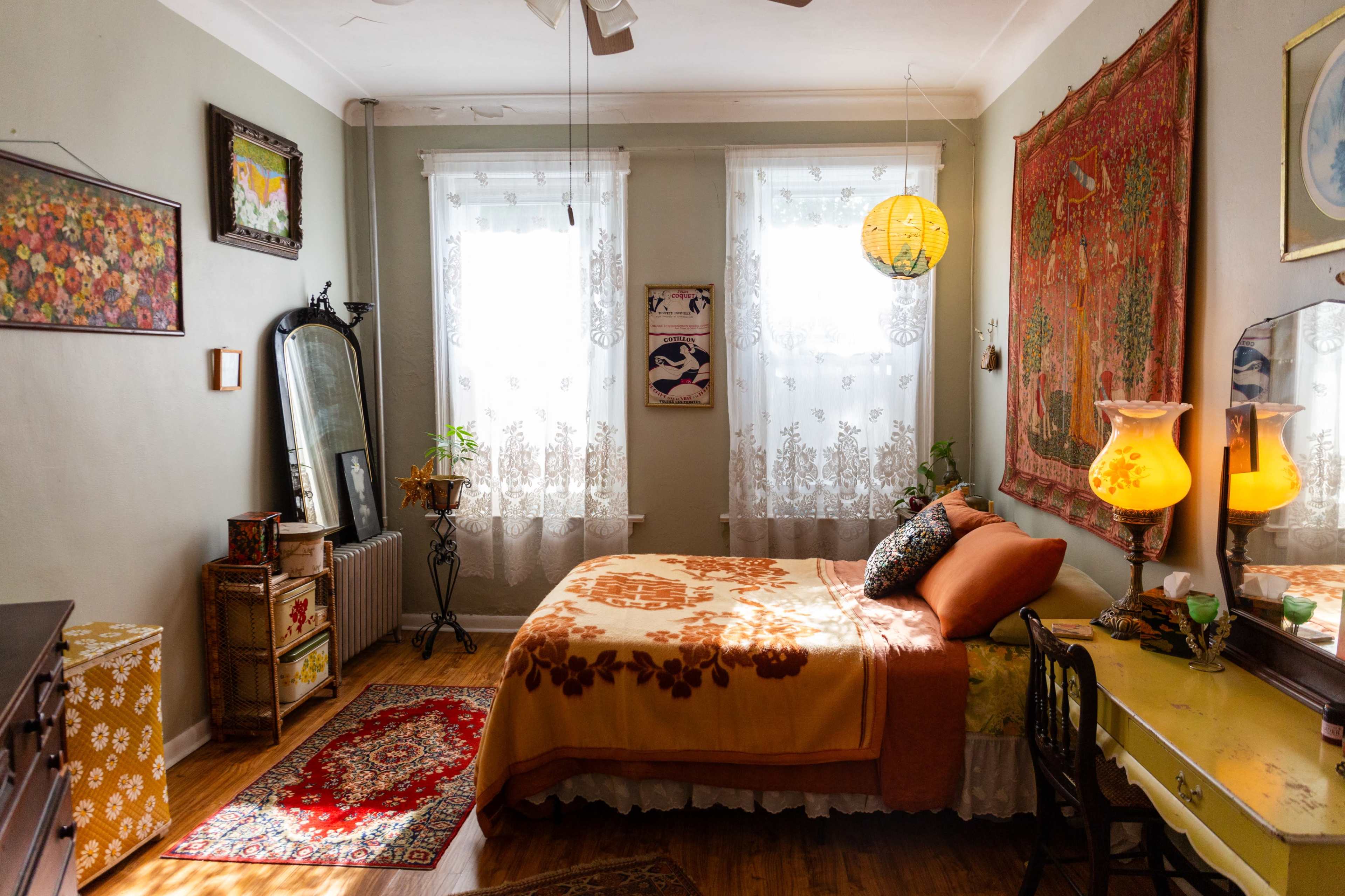 The image shows a cozy bedroom featuring a bed with an orange and white cover, a dresser, a vintage mirror, and large windows adorned with lace curtains.