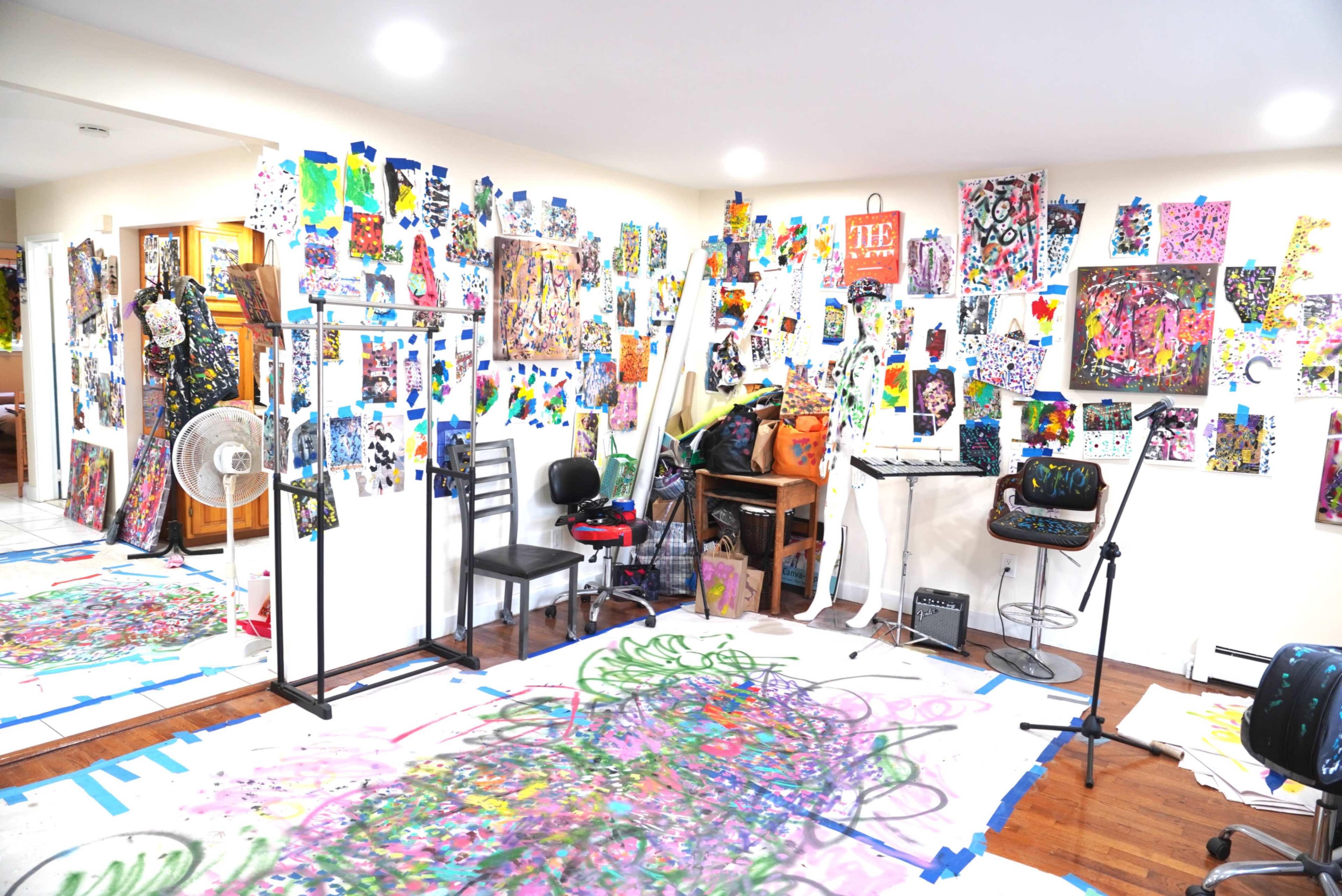 The image shows a vibrant art studio filled with colorful paintings and artworks displayed on walls and the floor.