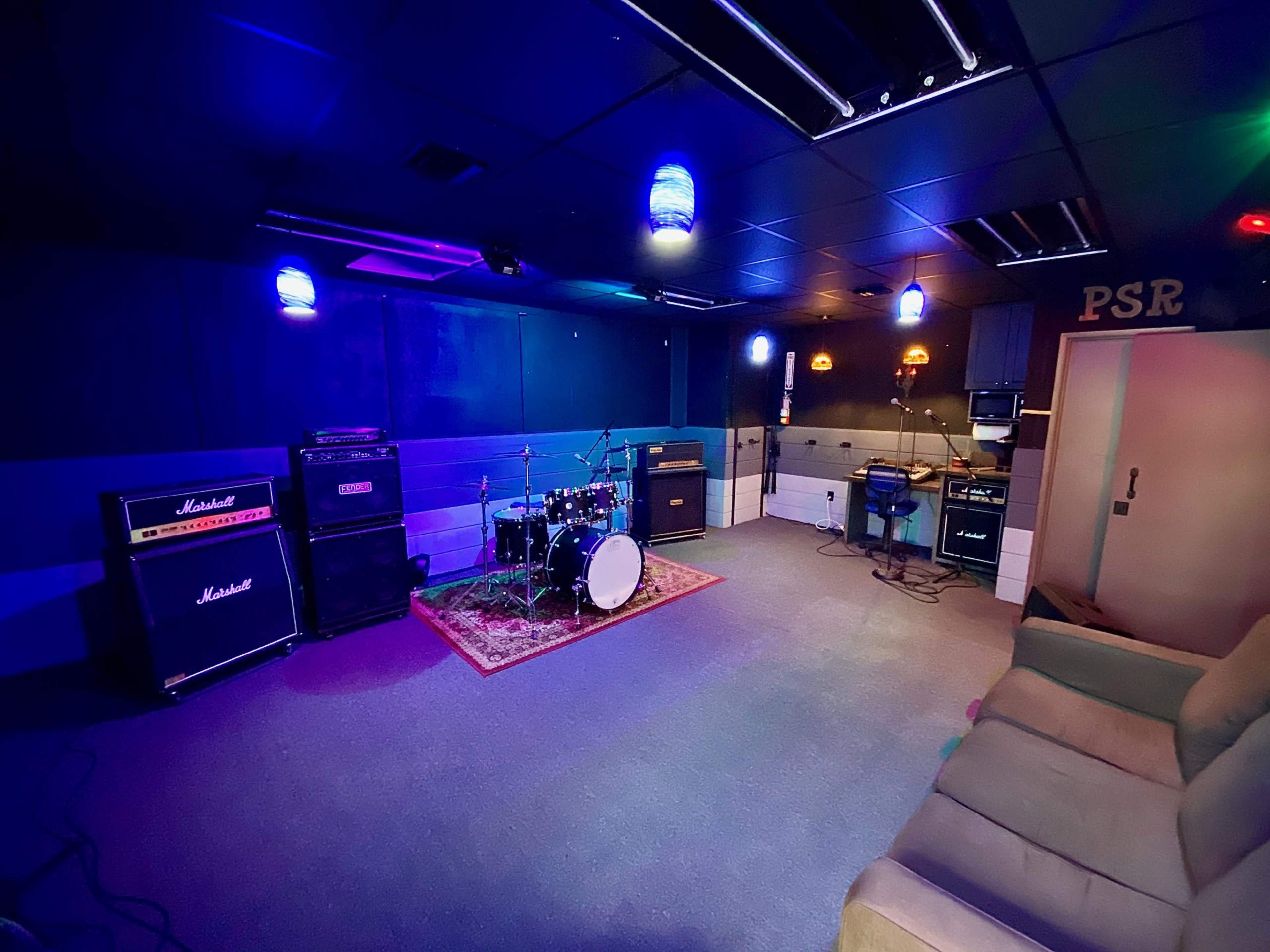 A music rehearsal room featuring drum equipment, amplifiers, and a couch, with colorful overhead lighting.