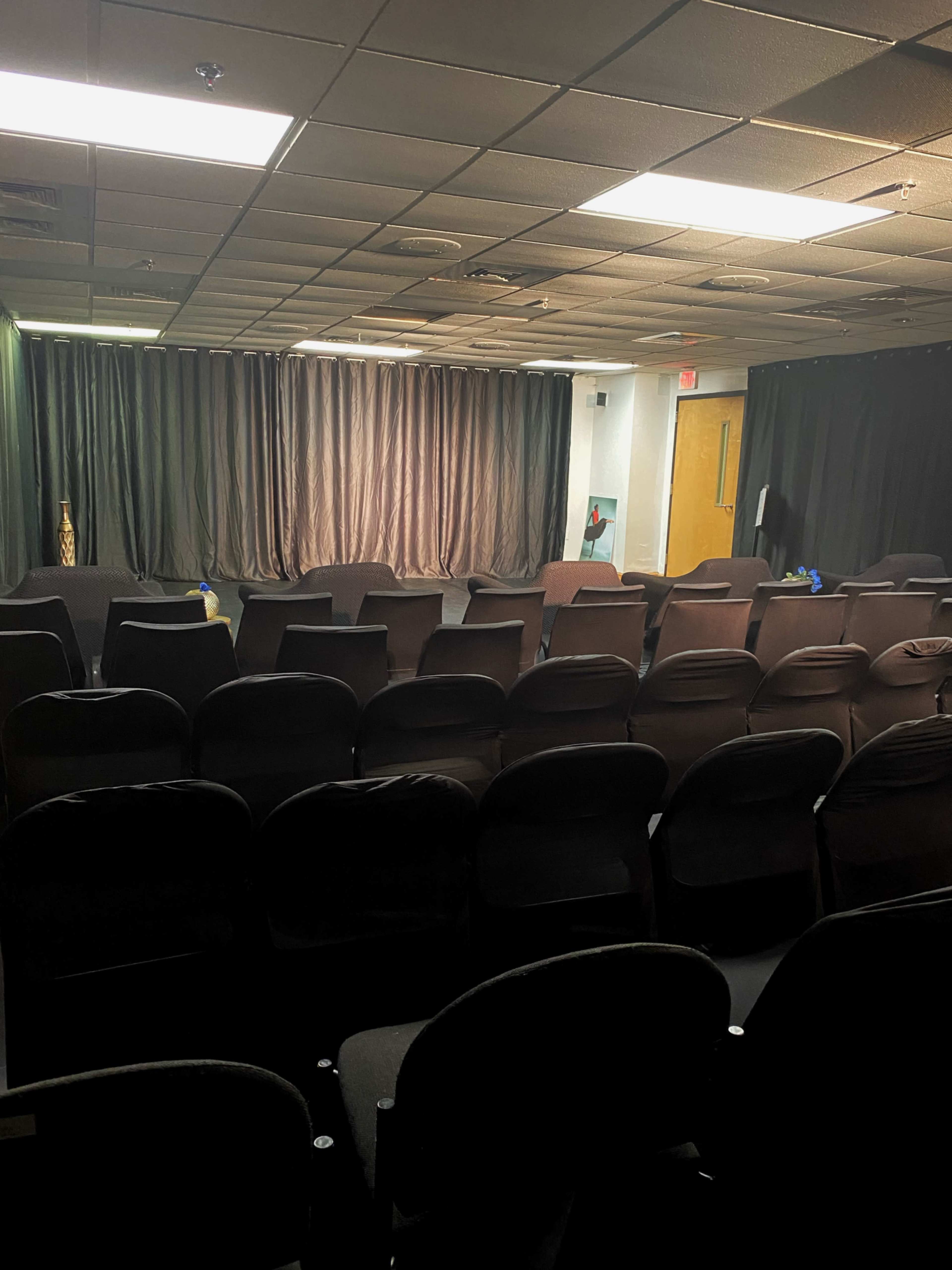 A small theater space features rows of black chairs facing a stage with black curtains.