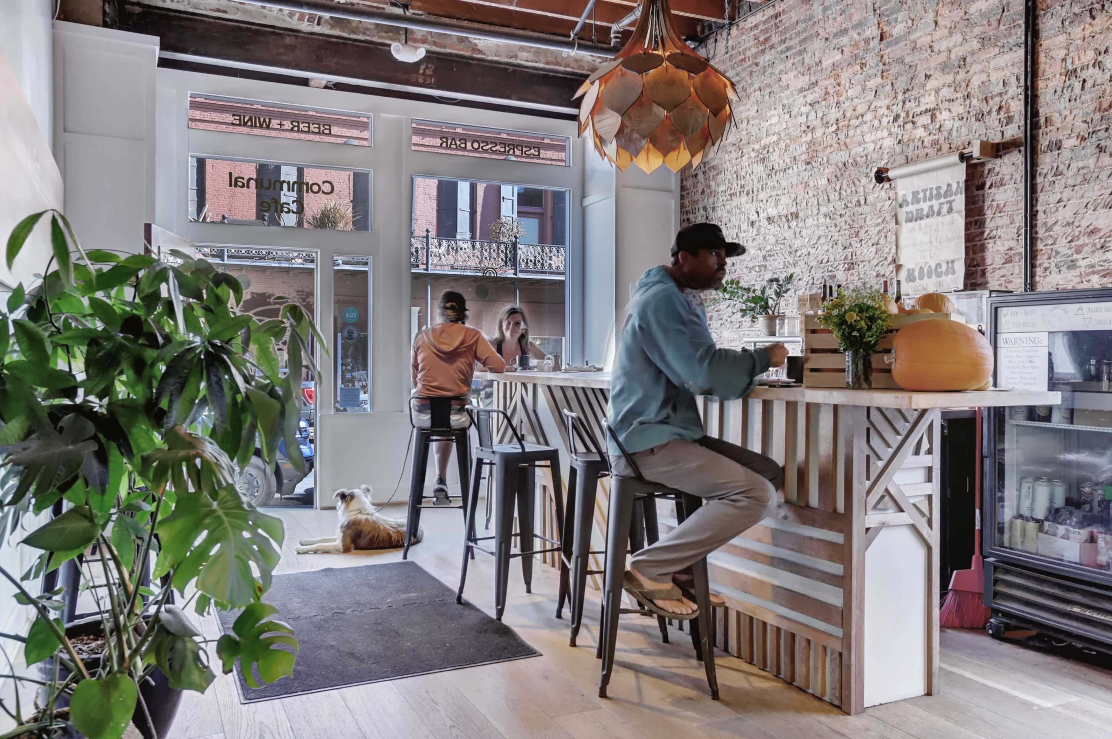 A man sits at a bar in a rustic cafe while two customers are seen at a table in the background, and a dog rests on the floor nearby.