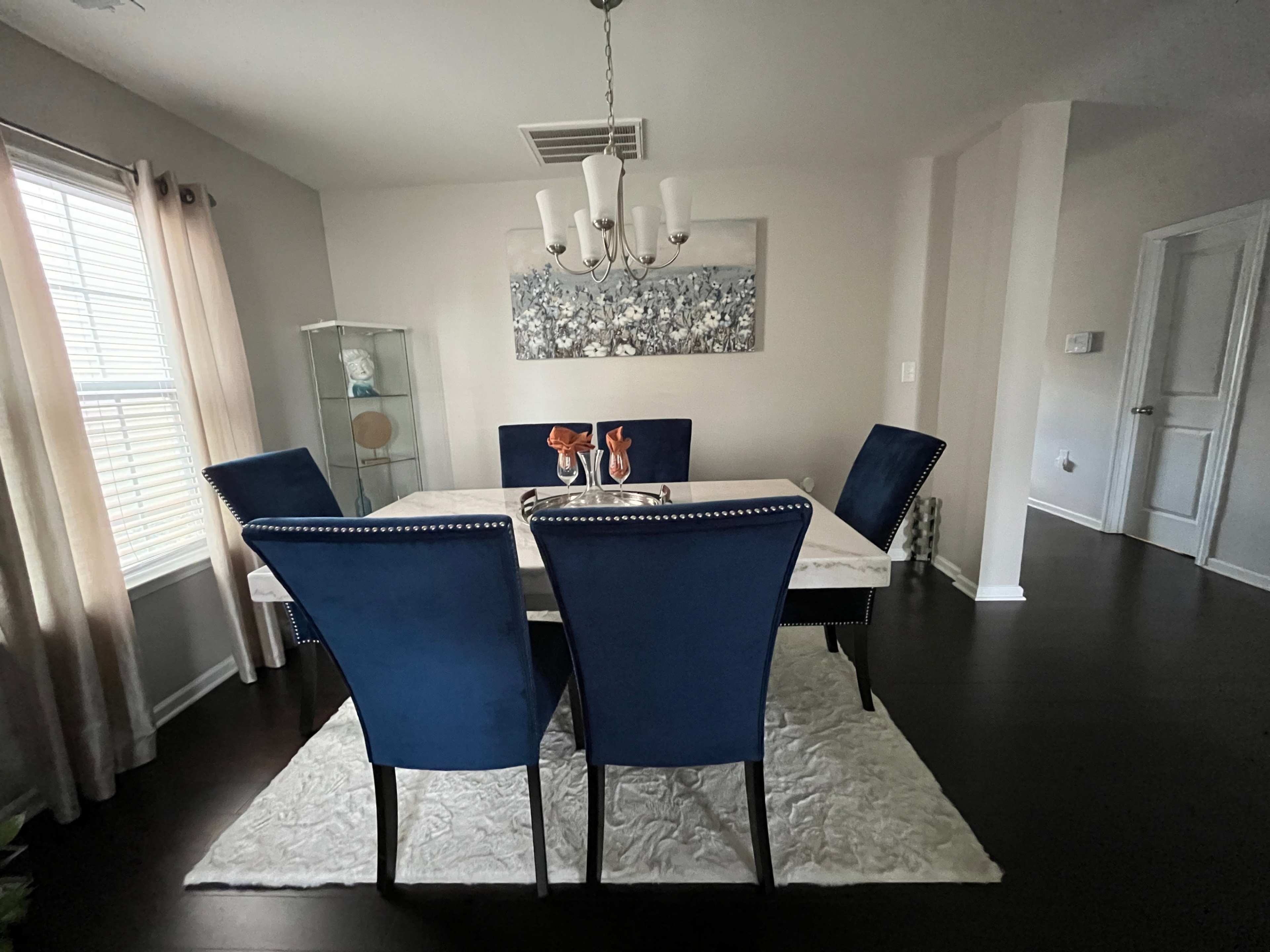 A dining room features a white table surrounded by four blue chairs, with a chandelier overhead and a decorative piece on the wall.
