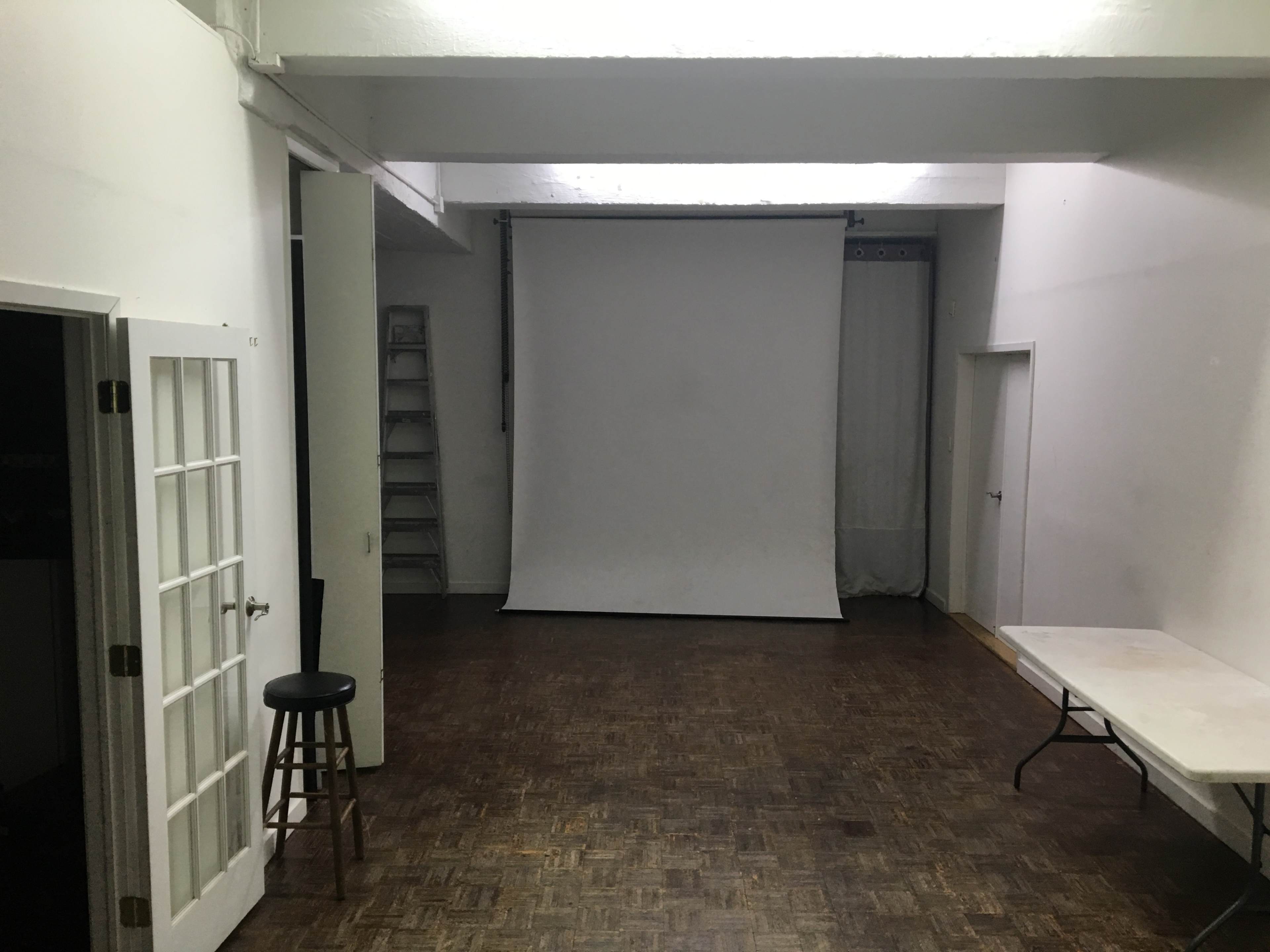 A spacious, empty room features a white backdrop on a stand, a folding table, a stool, and a ladder against the wall.